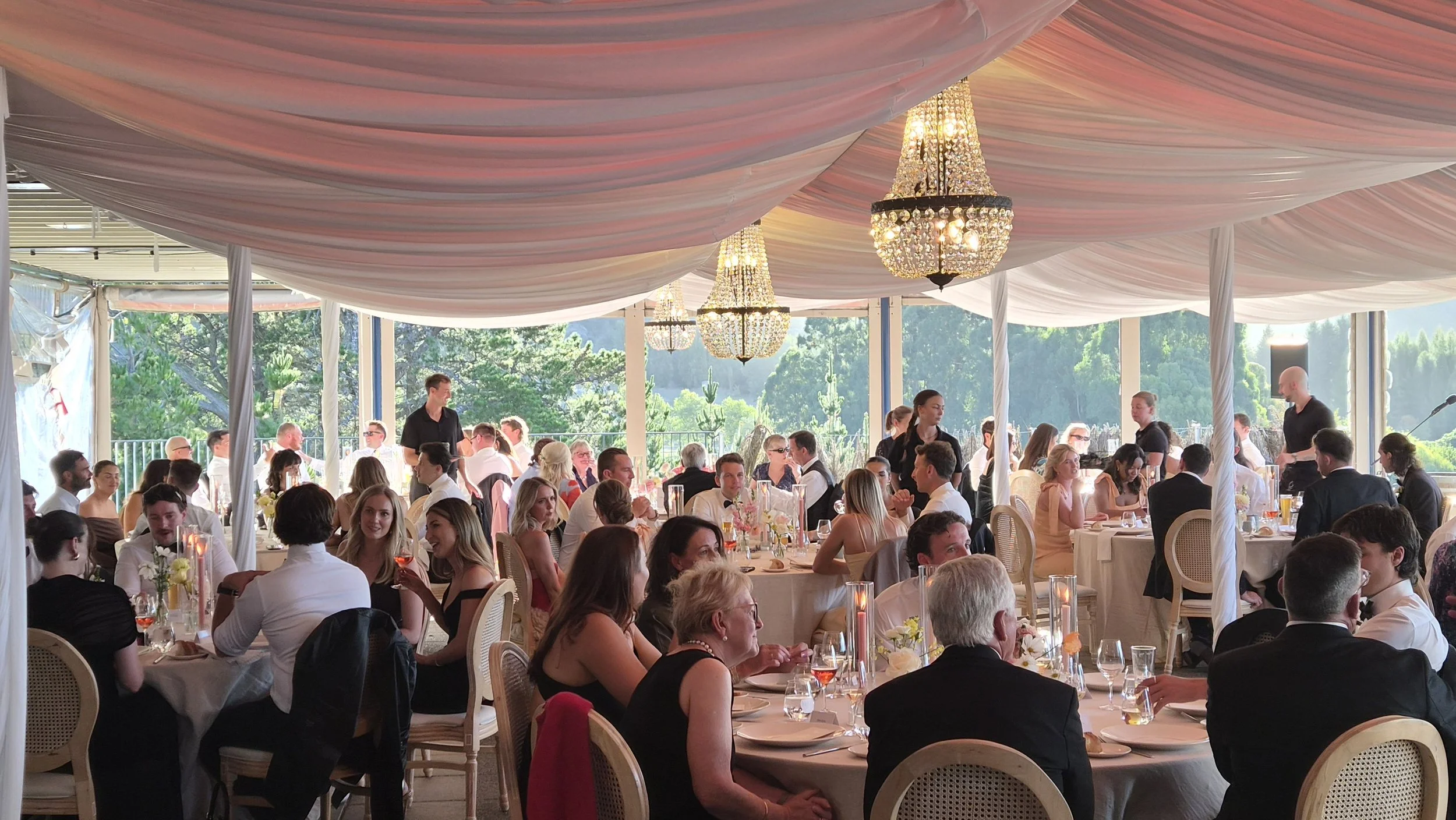 A large indoor wedding reception with many guests seated at decorated tables, chandeliers hanging from a draped pink ceiling, and large windows showing greenery outside.