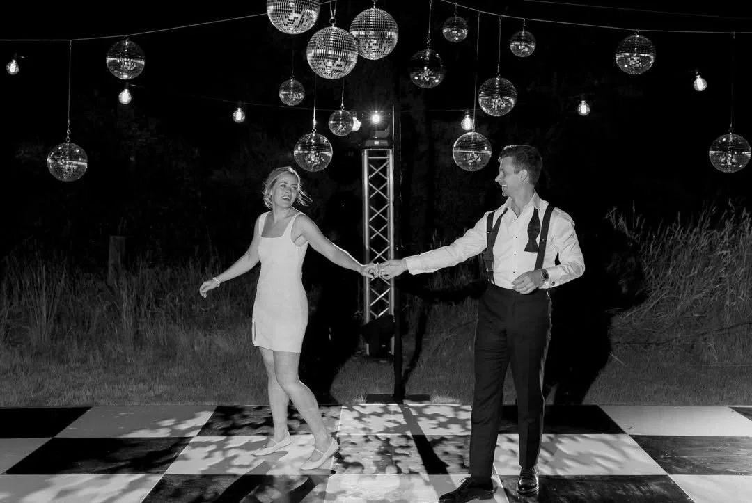A couple dancing at night under hanging disco balls and string lights, backlit by a bright light.