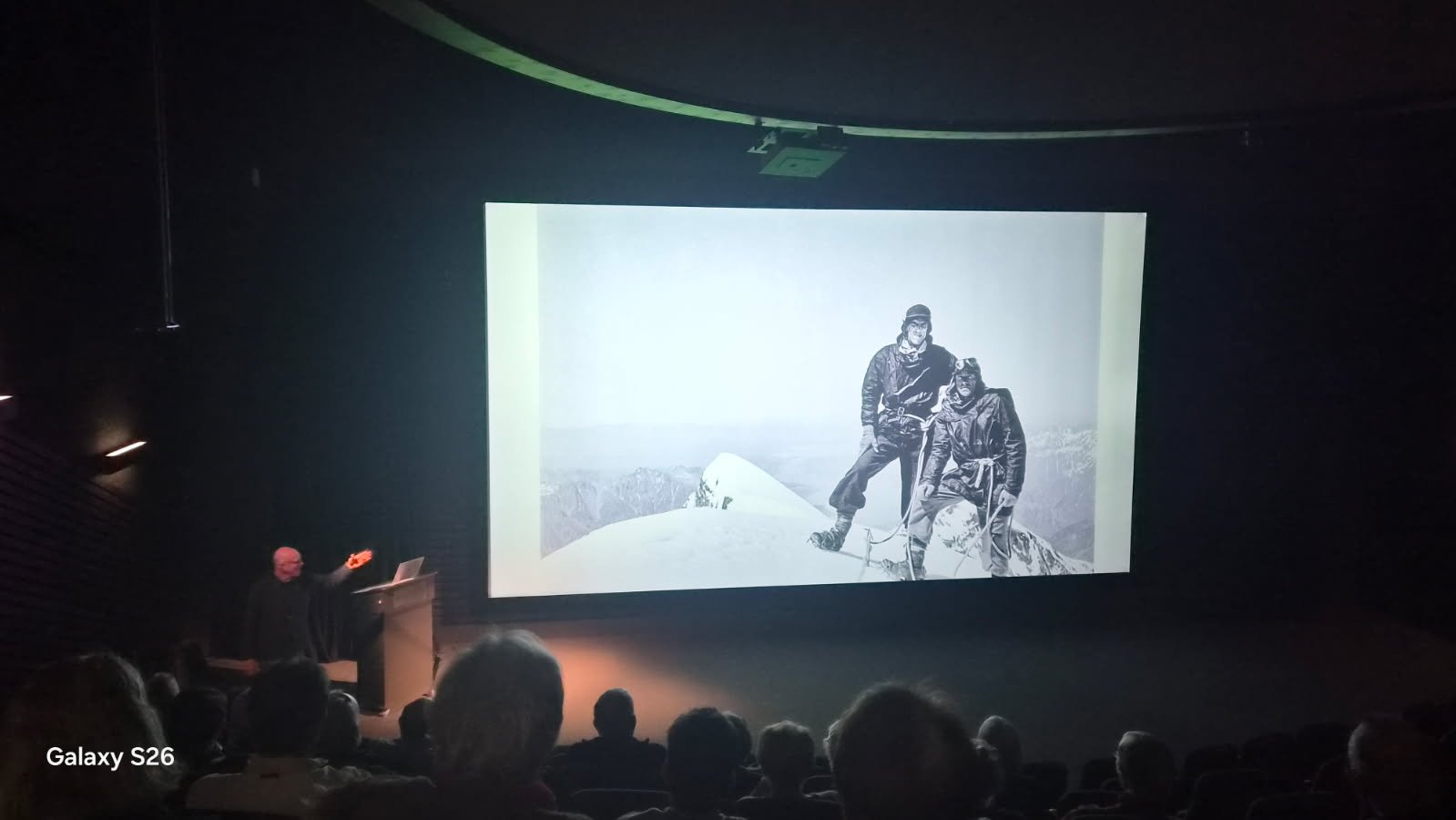 A black-and-white photograph displayed on a large screen showing two mountaineers on a snowy mountain peak with mountain ranges in the background.