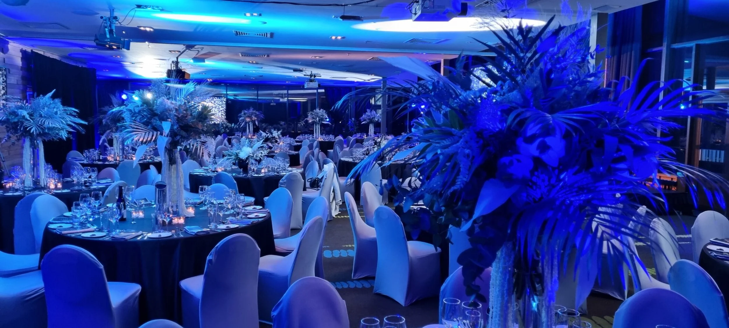 A banquet hall decorated for a formal event with round tables covered in dark blue tablecloths, white chairs, and tall floral centerpieces illuminated with blue lighting, creating an elegant atmosphere.