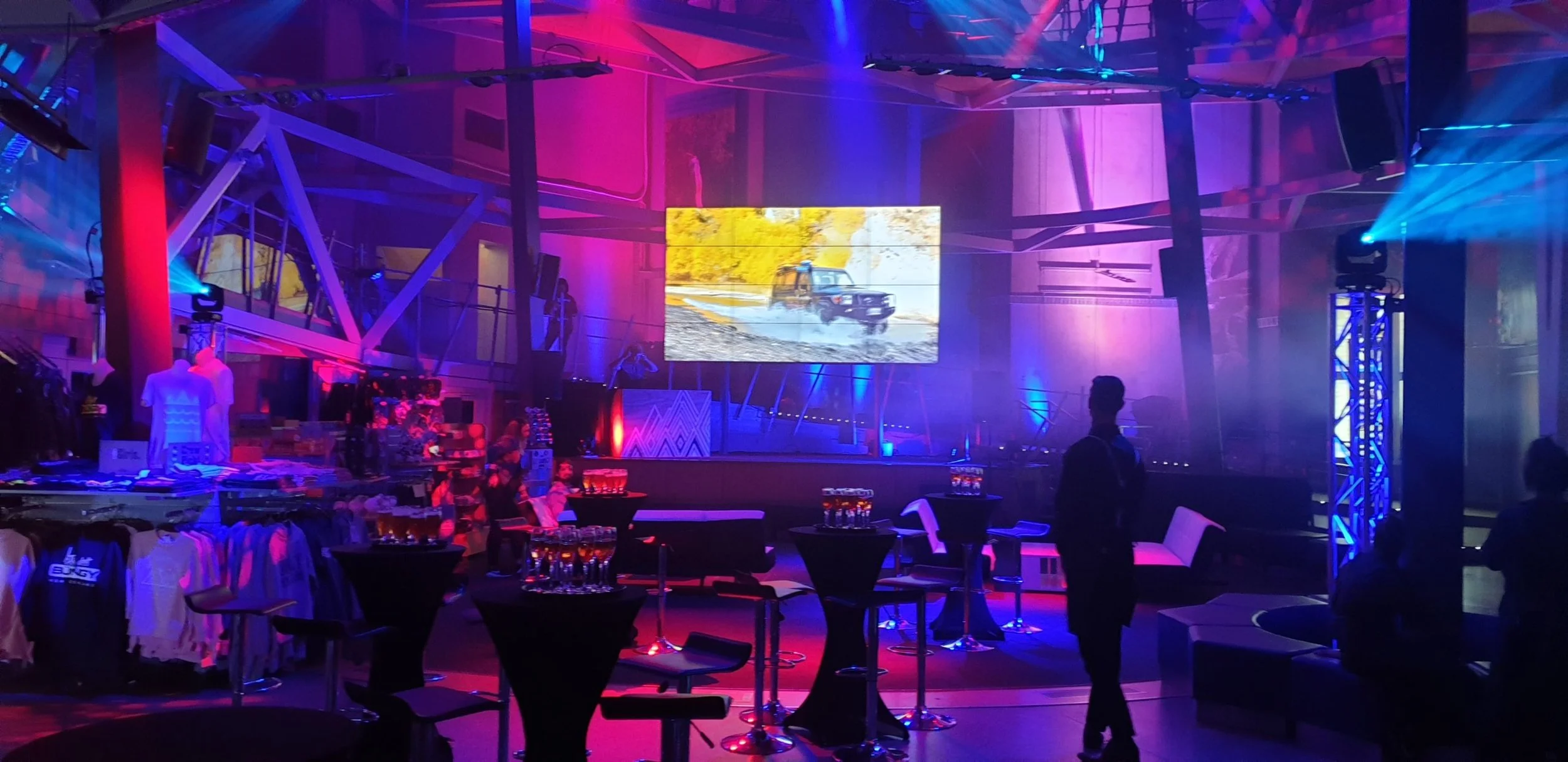 A dimly lit event space with colorful purple, blue, and pink lighting, a large screen displaying a vehicle driving through a muddy terrain, and tables with drinks and merchandise, suggesting a party or presentation with a tech or automotive theme.