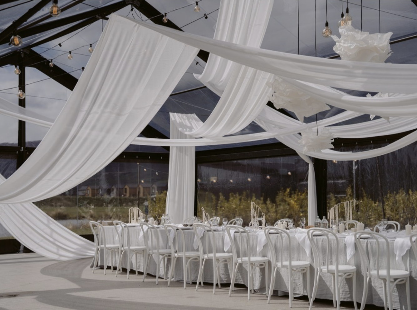 Decorated event space with white draped fabric flowing from the ceiling and a long banquet table set with white chairs, glassware, and decorative centerpieces, enclosed by glass walls revealing outdoor scenery.