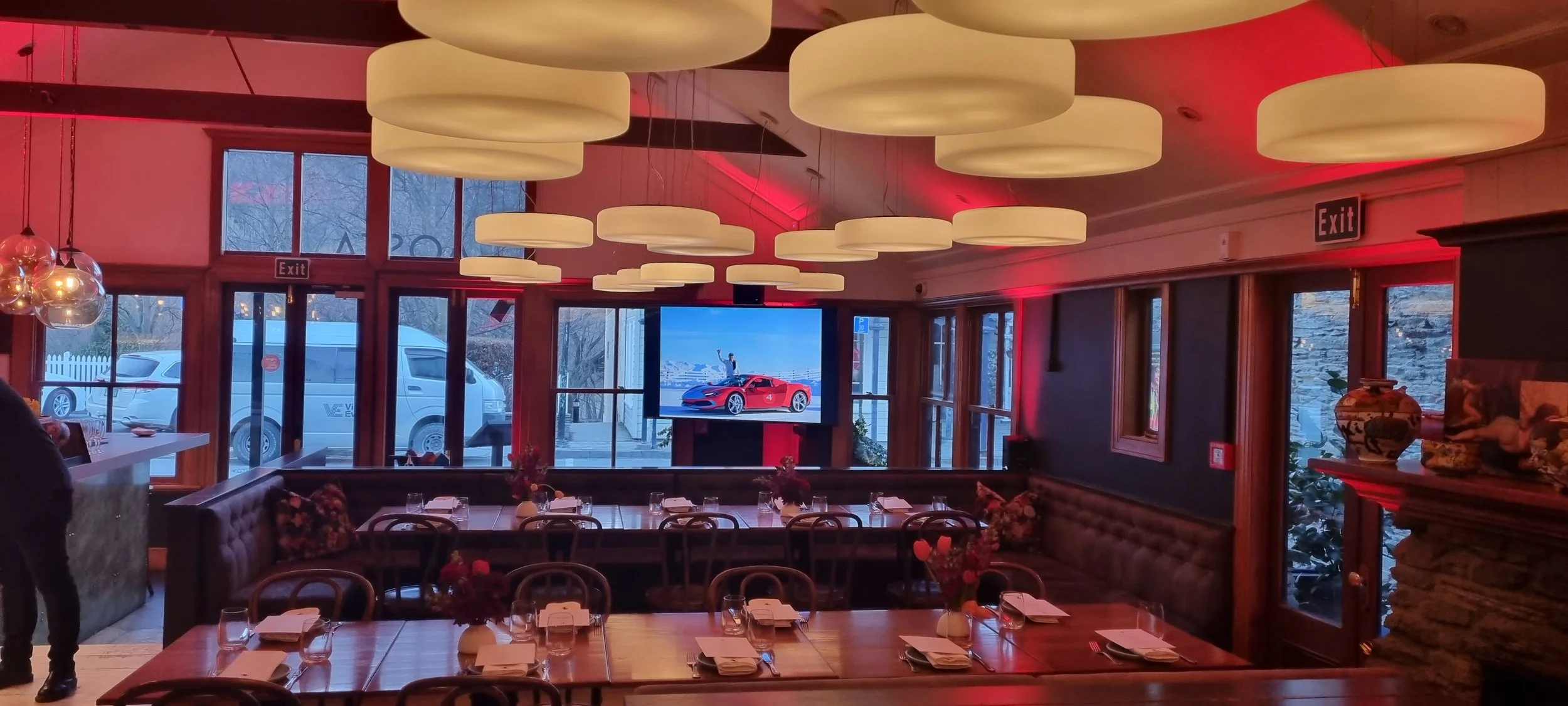 Interior of a restaurant with wooden tables set with napkins and glasses, decorated with small flower arrangements. Large, off-white, circular pendant lights hang from the ceiling. There are windows and glass doors with views of parked vehicles outside. A large television screen on the wall displays a red sports car and a person waving. Red accent lighting is present in the room.
