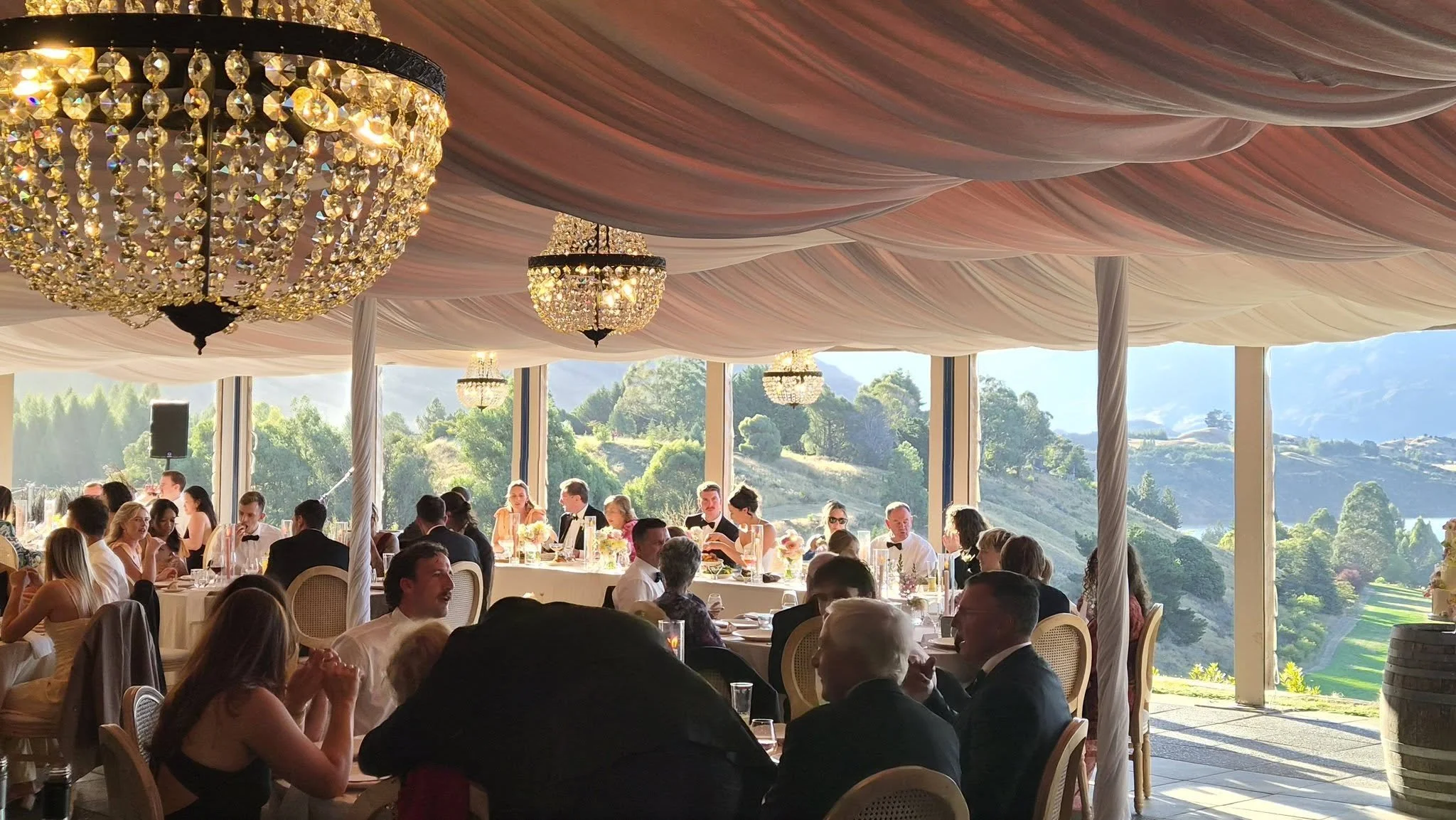 People dressed in formal attire sitting at round tables with white tablecloths, in a large event space decorated with pink draped fabric ceiling and crystal chandeliers, with large windows revealing a scenic outdoor landscape of green trees and hills.