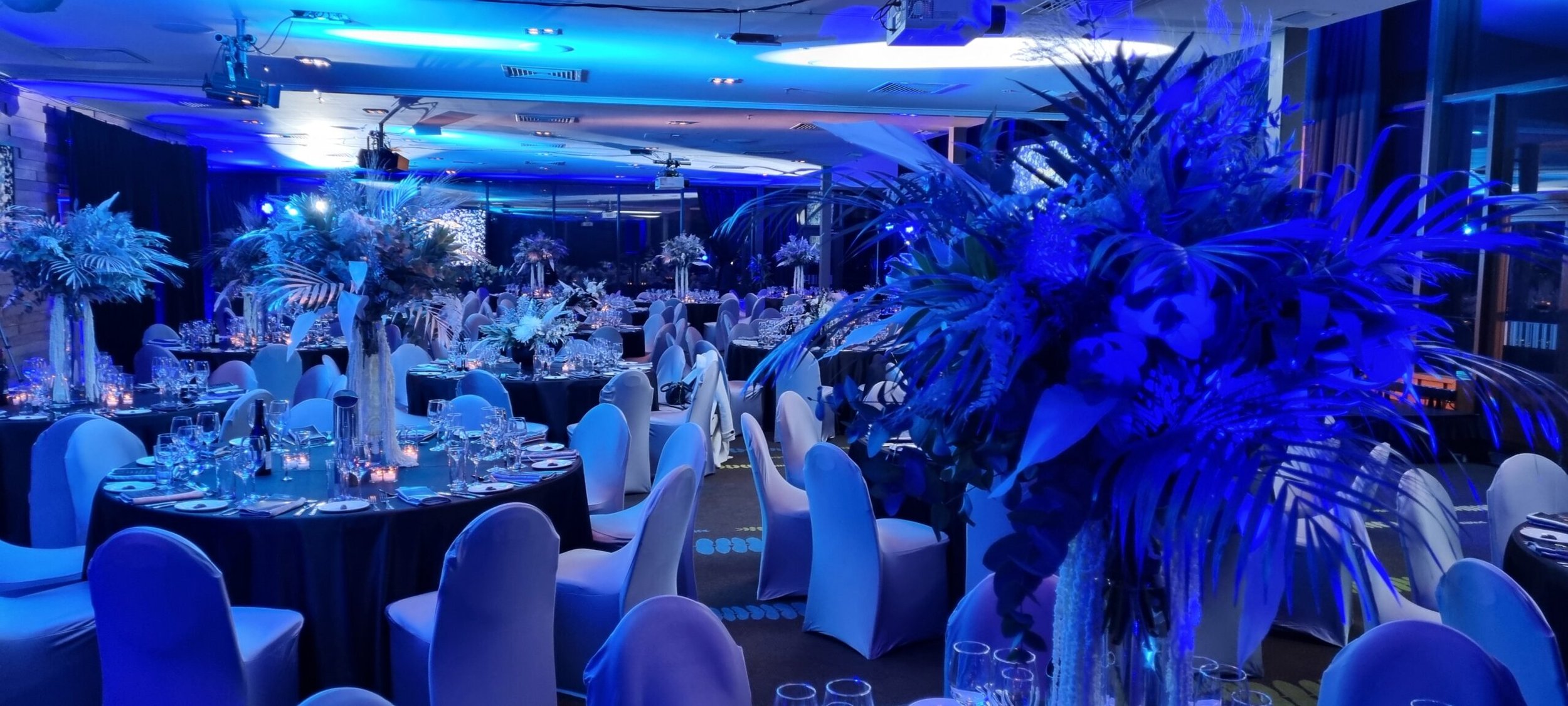 An indoor event space decorated with floral centerpieces and blue lighting, possibly set up for a wedding or banquet.