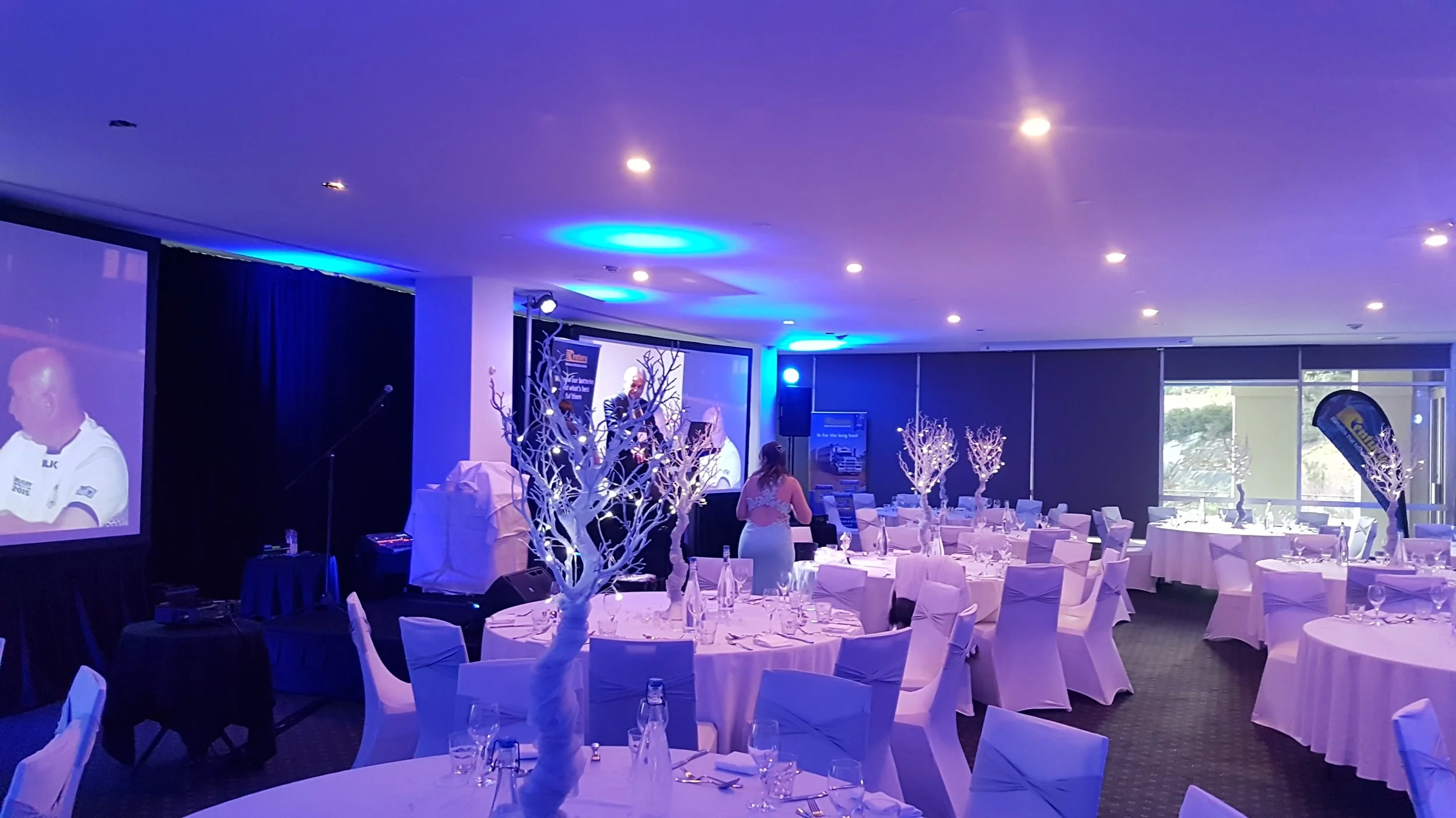 Indoor banquet hall decorated for an event with round tables covered in white tablecloths and white chairs. Centerpieces are illuminated white tree branches. A stage with a speaker and a large screen displaying a man in a white uniform and a blue ban