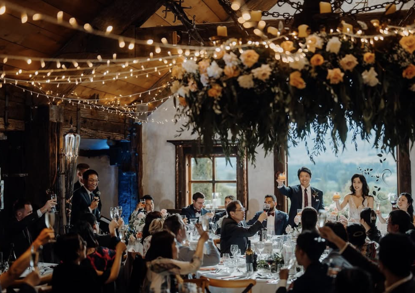 A wedding reception in a rustic barn with string lights and floral decorations, where guests are raising glasses in a toast as the bride and groom smile.