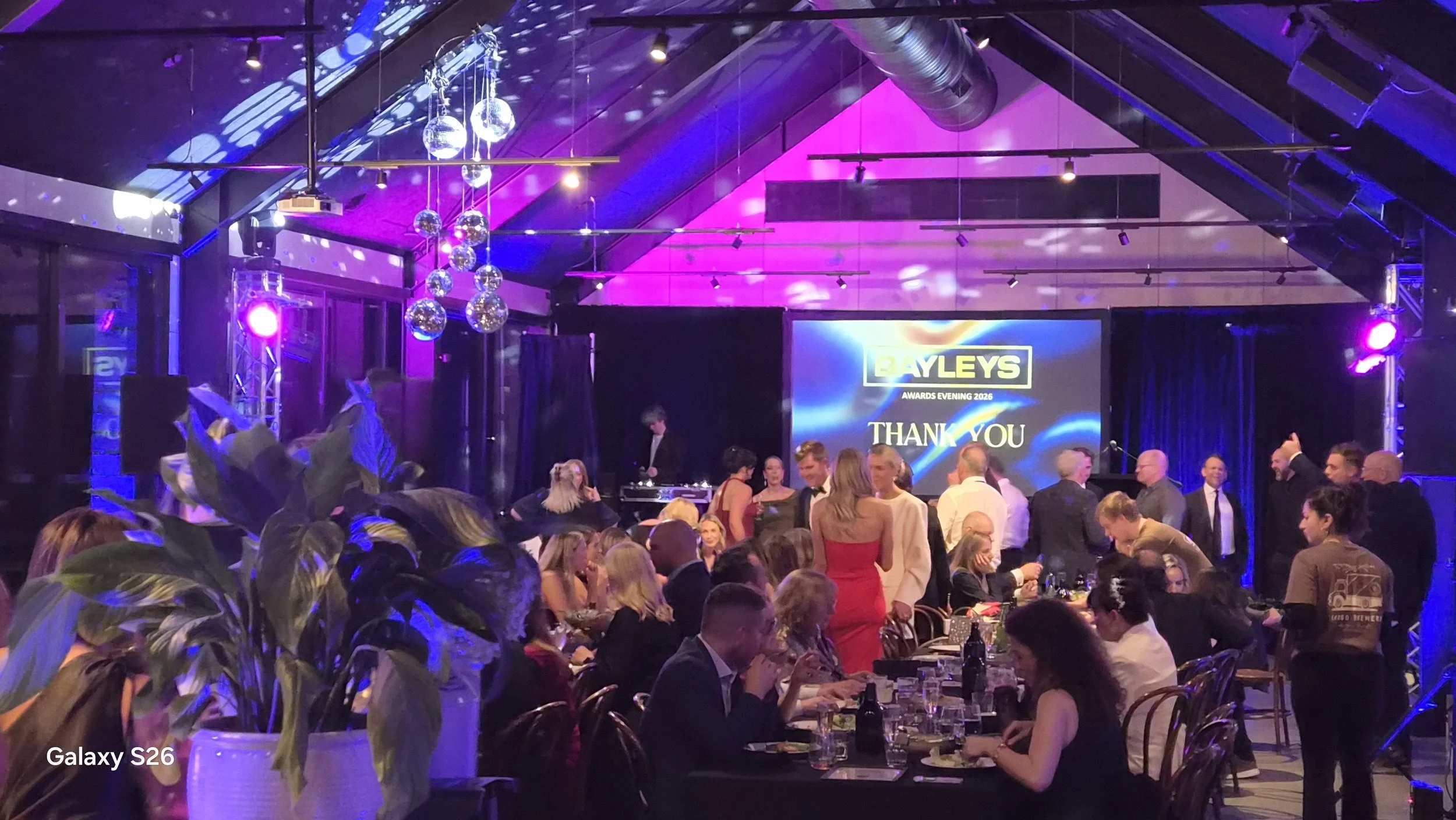 A lively indoor awards event with people dressed in formal attire, a large screen displaying 'Bayleys Awards Evening 2026' and 'Thank You,' decorated with purple and blue lighting, hanging disco balls, and a stage with a DJ.