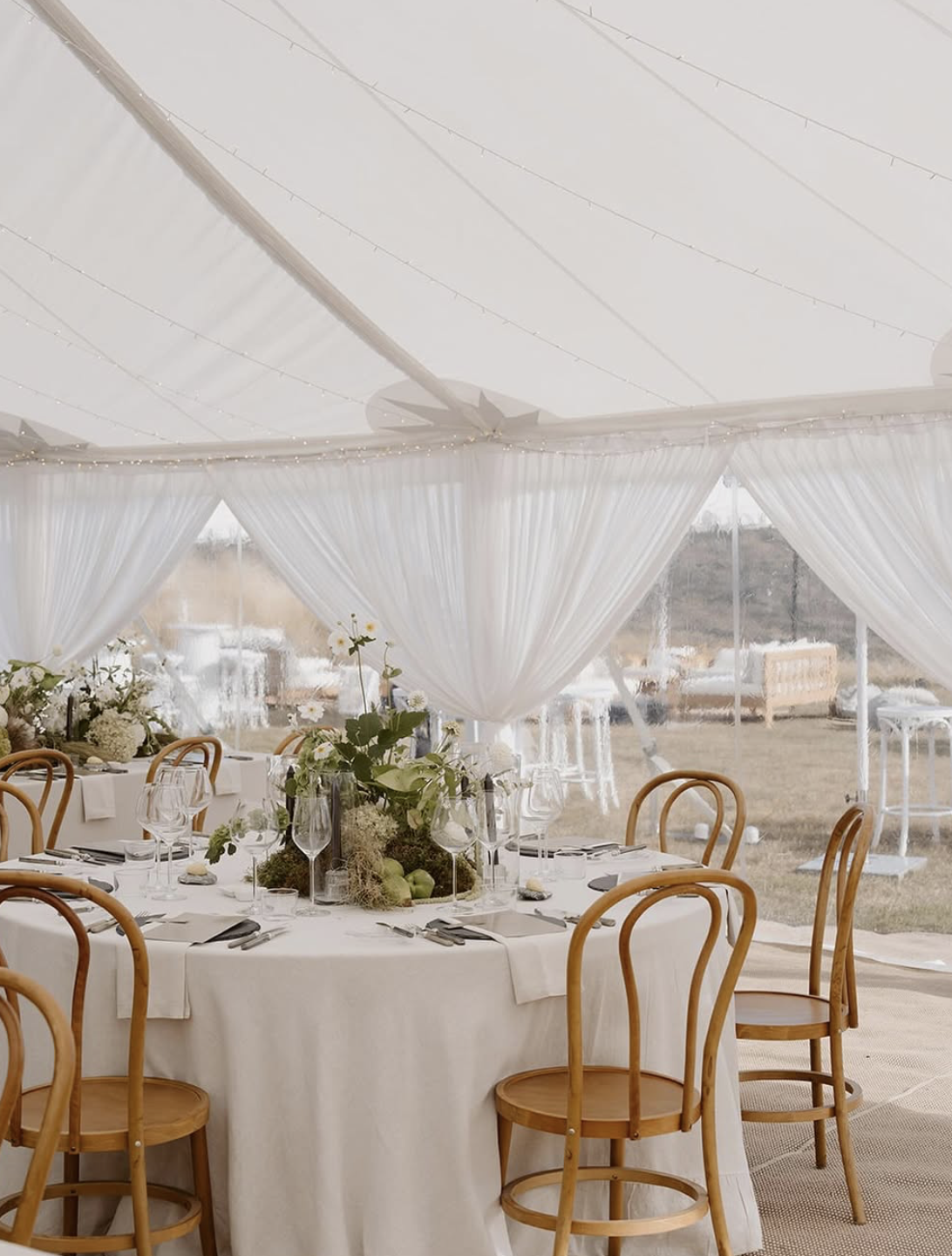 Elegant event tent with white drapery, decorated table with floral centerpiece, glassware, and wooden chairs.