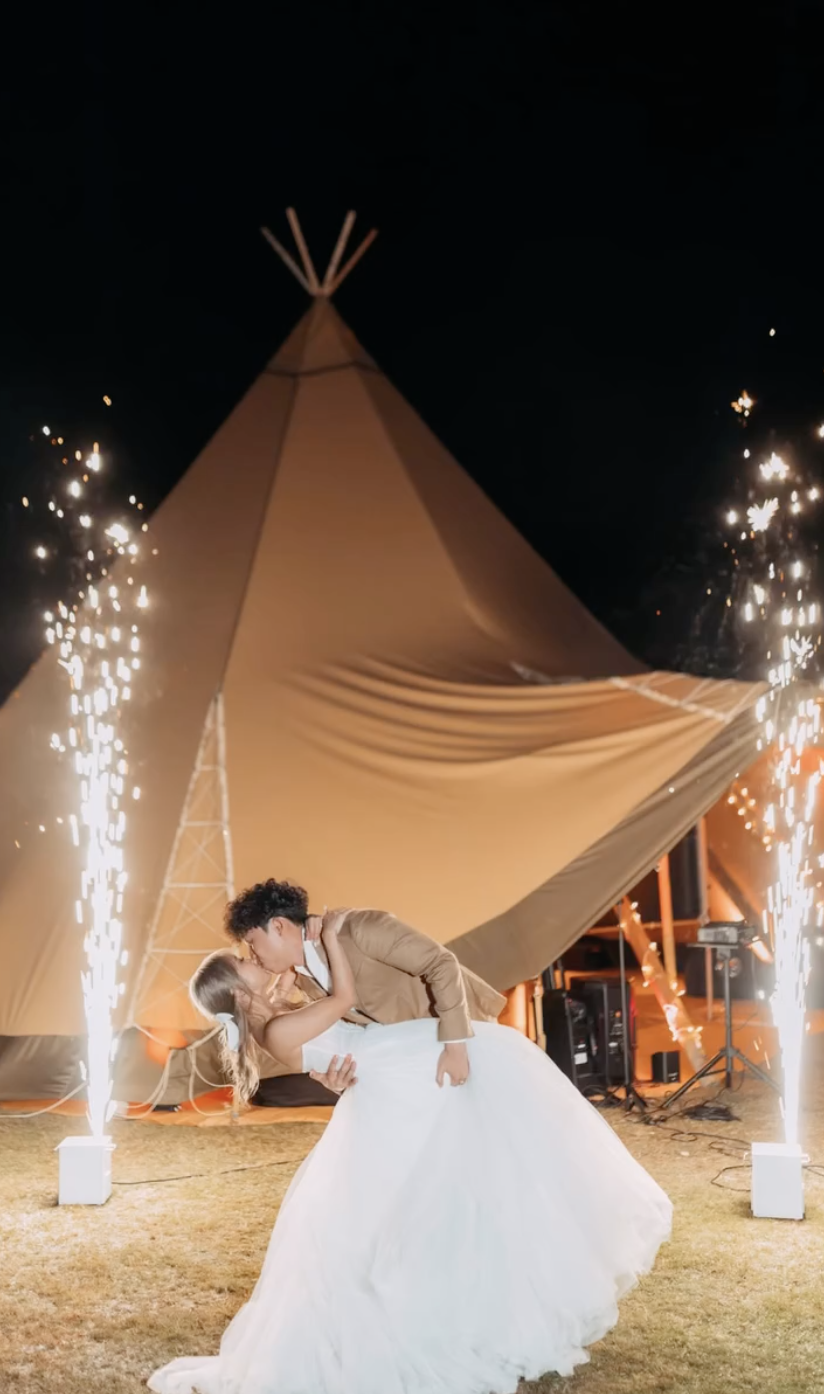 A newlywed couple sharing a kiss at night during their wedding celebration, with sparks and fireworks in the background and a large tent behind them.