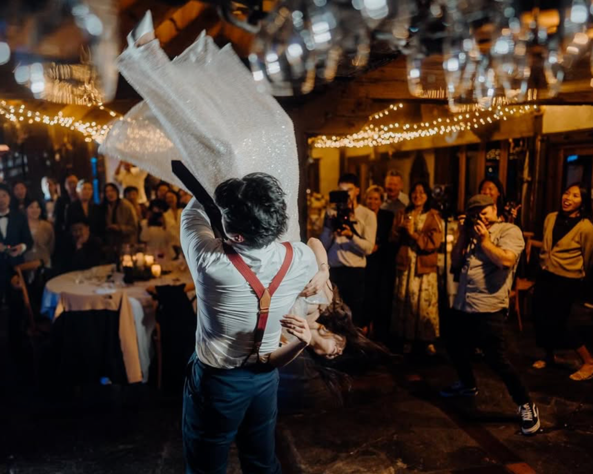 A man in suspenders twirling a woman in a white dress at a lively indoor celebration with guests watching and taking photos, decorated with string lights.