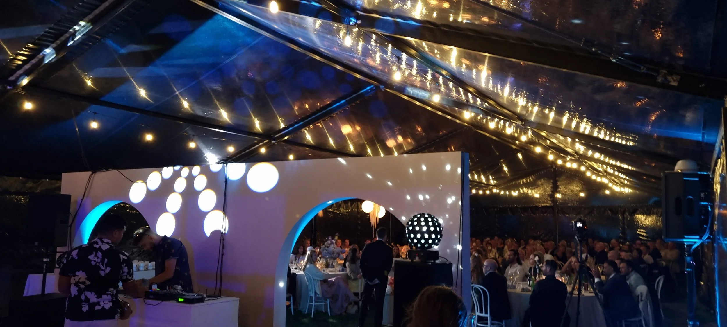 A nighttime event under a large reflective tent lit with warm string lights. Inside, many people are seated at tables, and there is a DJ booth with two people working. The tent has a modern design with circular cutouts and polka dot lights.