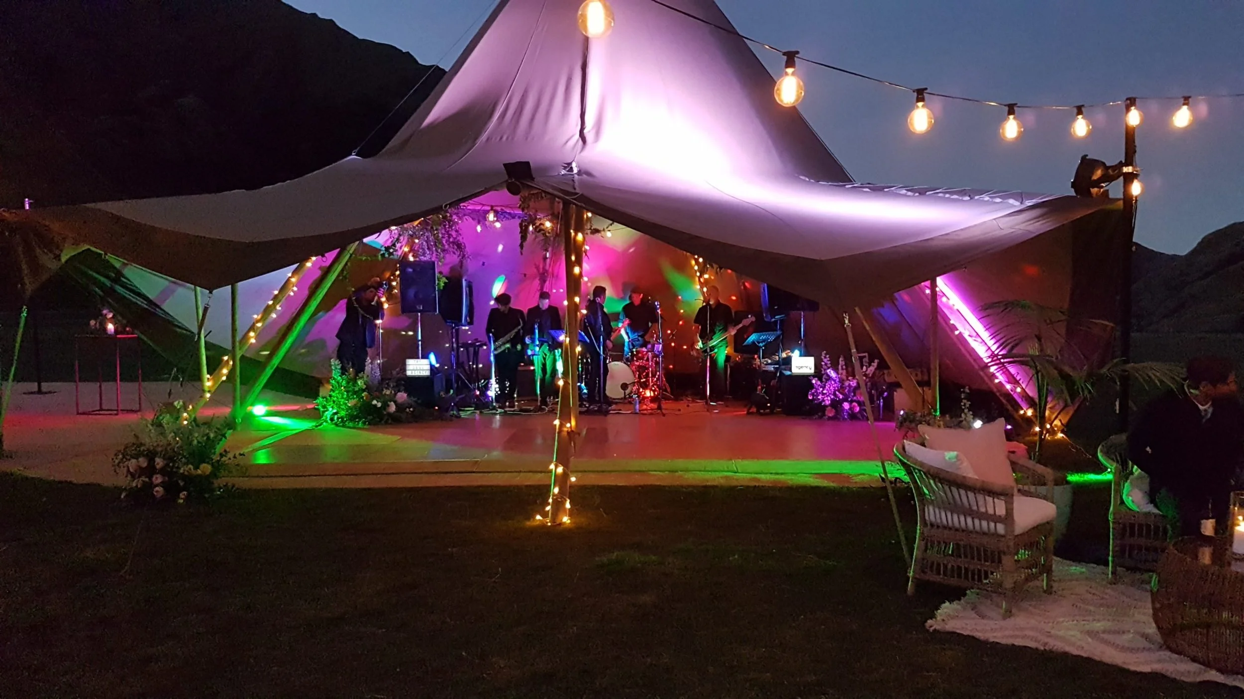 Outdoor night scene featuring a stage with a band playing under a large canopy, decorated with string lights and colorful lighting effects, surrounded by floral arrangements and seating.