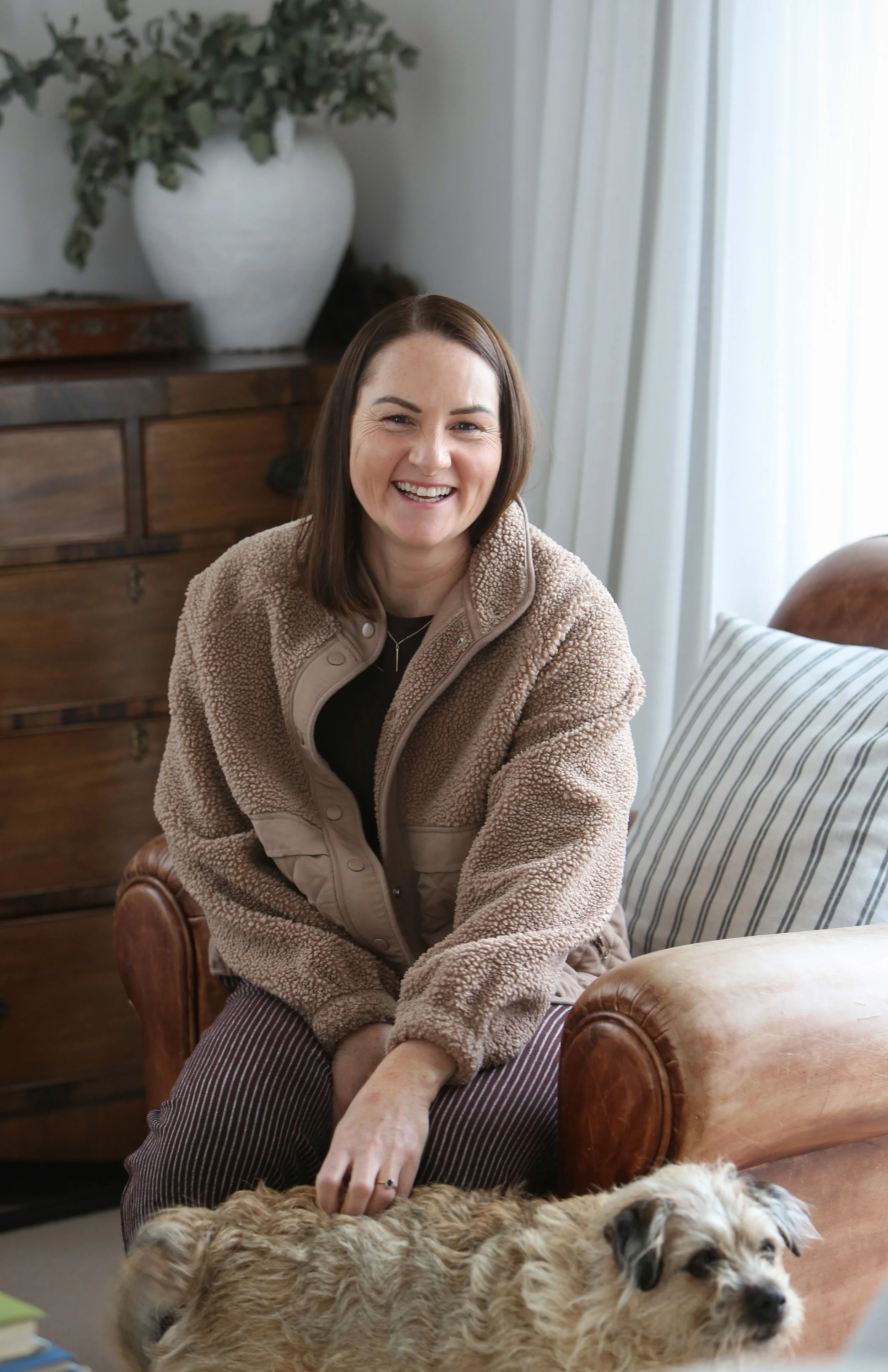 A woman with brown hair smiling while sitting in a living room with a small dog lying on the floor in front of her and a larger dog lying on a brown leather couch.