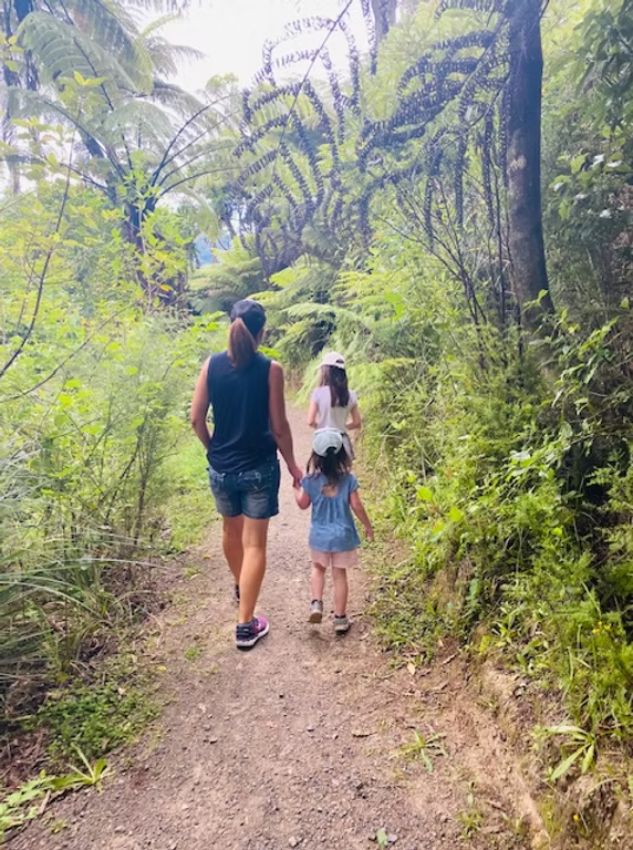 A woman and two young girls walking hand-in-hand along a trail in a lush, green forest with tall trees and fern-covered foliage.