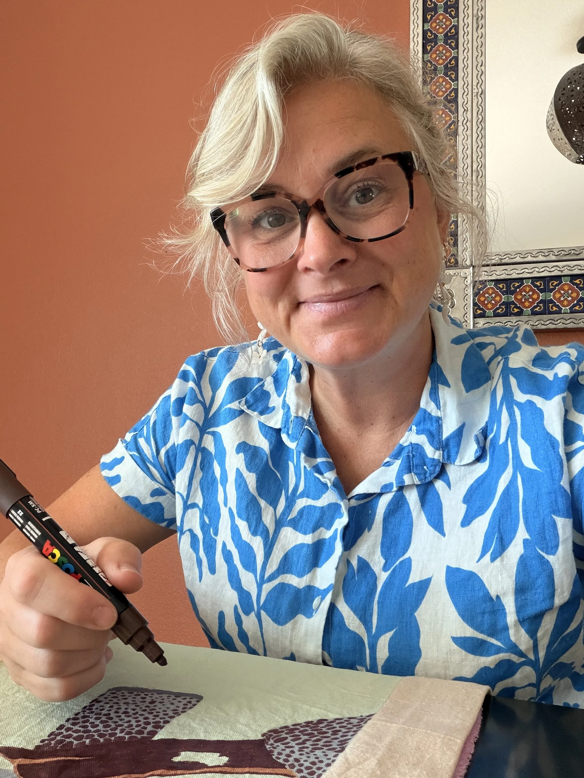 Woman with blonde hair and glasses, wearing a blue and white patterned shirt, sitting at a table and holding a black marker with colorful text on it. She is working on a piece of fabric or paper with a design of red, black, white, and green patterns. The background has an orange wall with decorative diamond-shaped artwork.