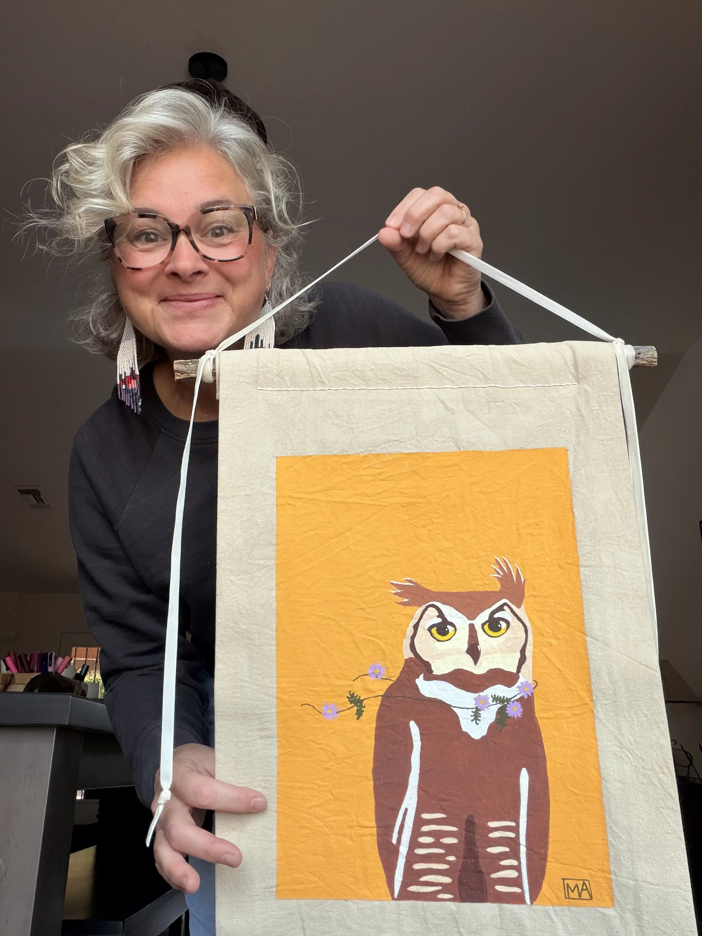 A woman with curly gray hair, glasses, and earrings holds a banner with an illustration of an owl on an orange background.