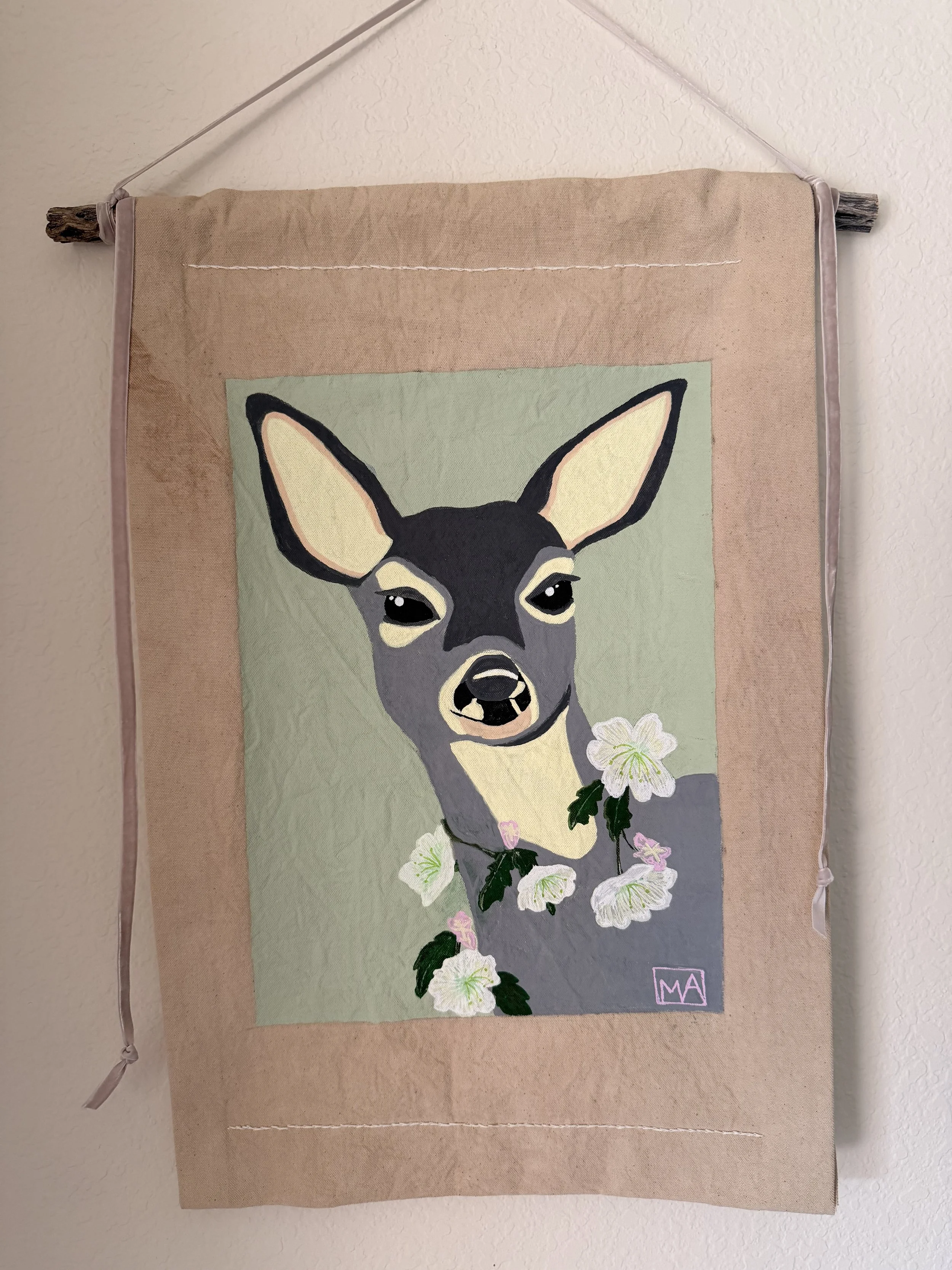 Wall hanging tapestry featuring a painted portrait of a deer with white flowers around its neck, hung on a beige wall.