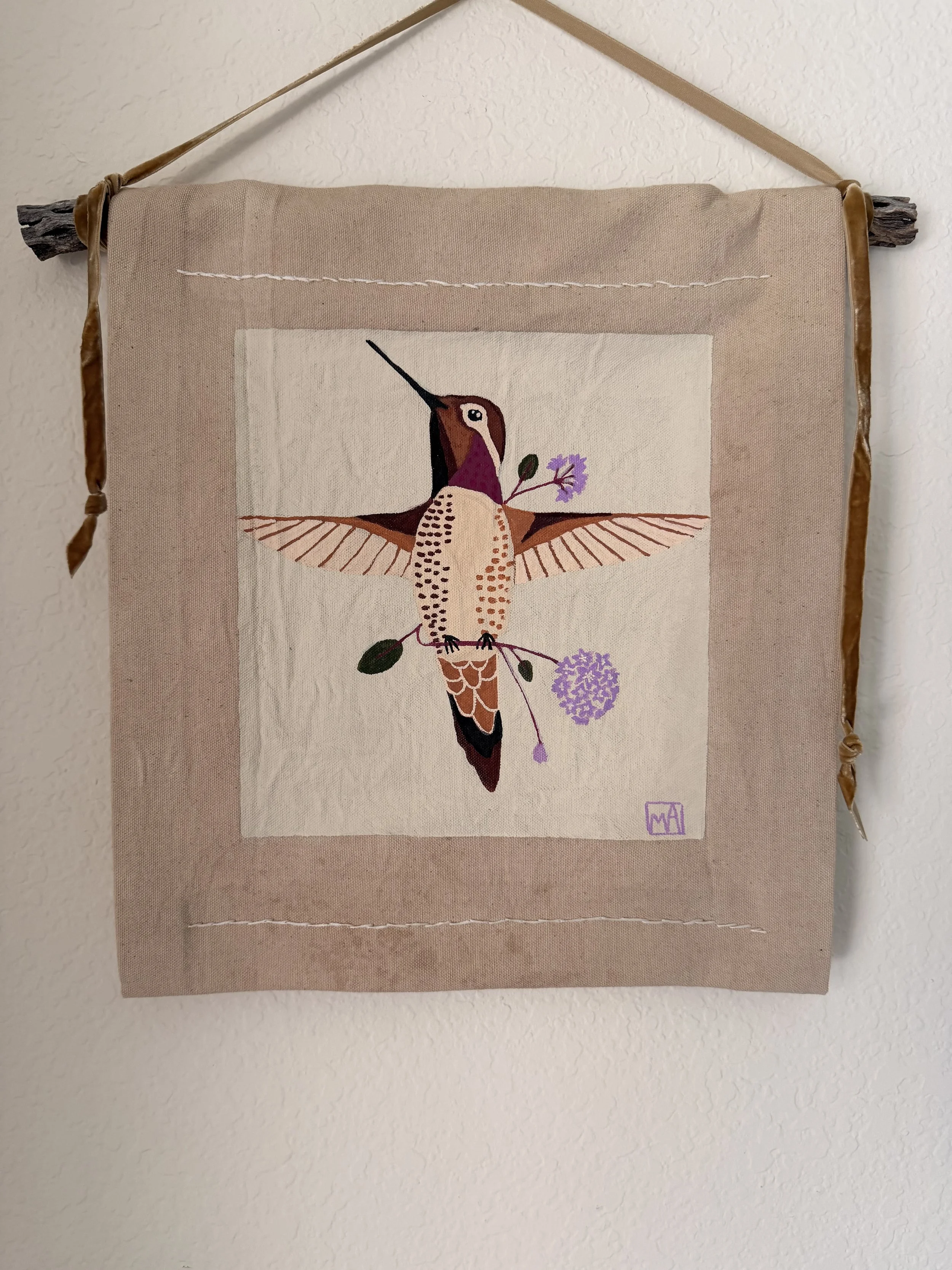 Wall hanging textile art featuring a stylized hummingbird holding purple flowers, mounted on a beige fabric background with a wooden rod at the top.