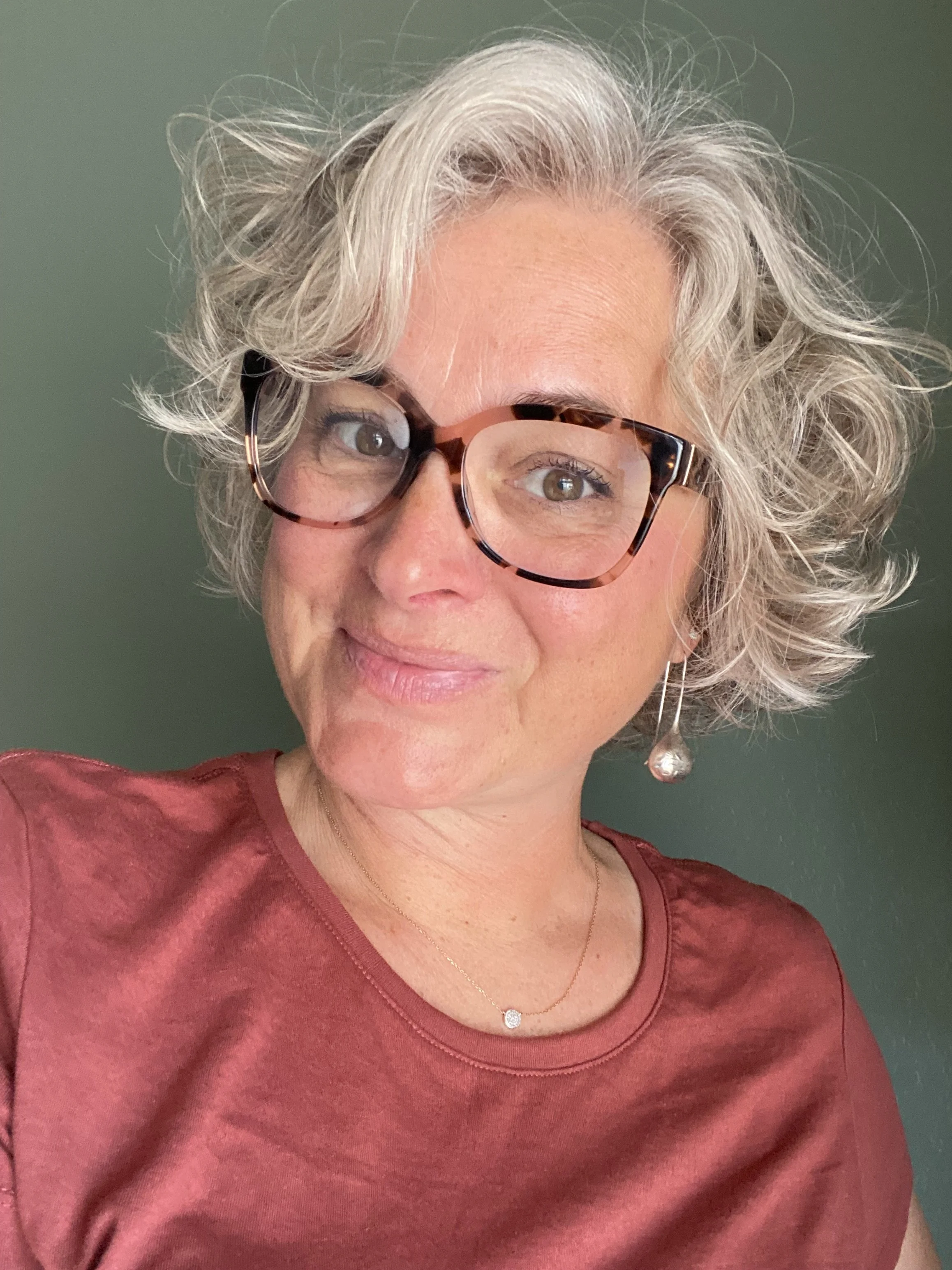 Close-up of a woman with gray, curly hair wearing eyeglasses, a red top, a small necklace with a pendant, and a teardrop-shaped earring, against a dark green background.