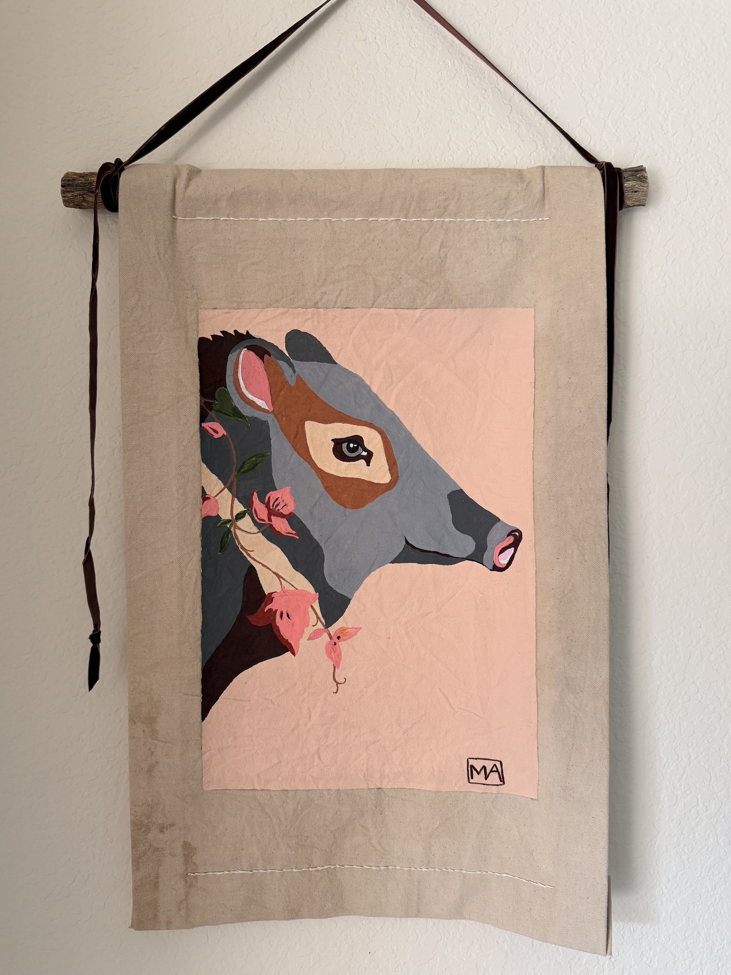 A wall hanging art piece featuring an illustrated horse's head with floral accents on a light pink background, displayed on a beige fabric banner with a wooden rod and ribbon for hanging.