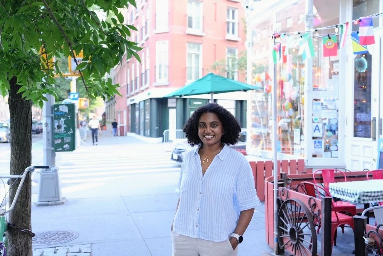 Sarya smiling in NYC with storefronts and outdoor seating.