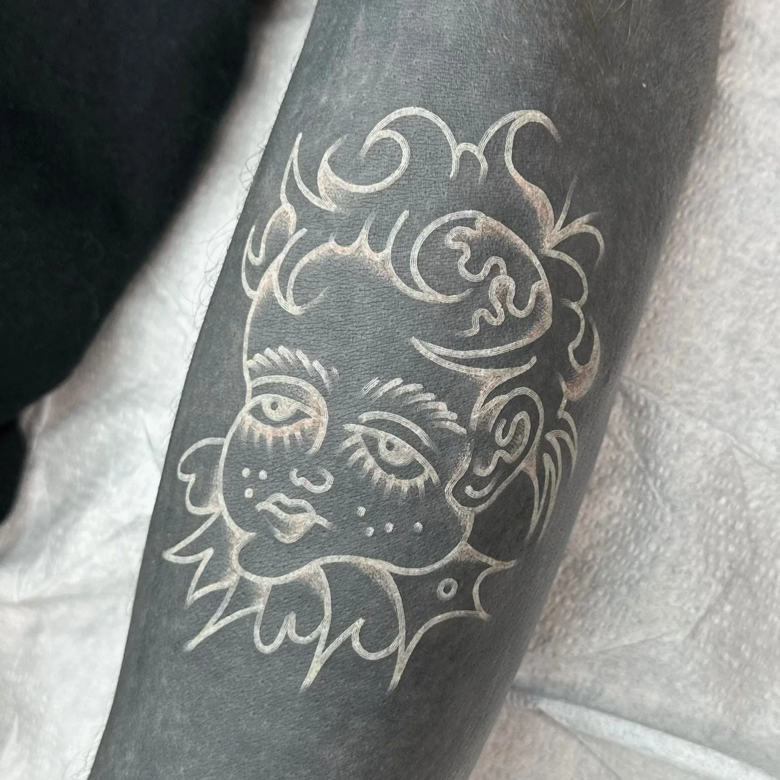 Close-up of a tattoo featuring a detailed, stylized portrait of a doll, white lines blasted over a black cover up, on a person's skin.