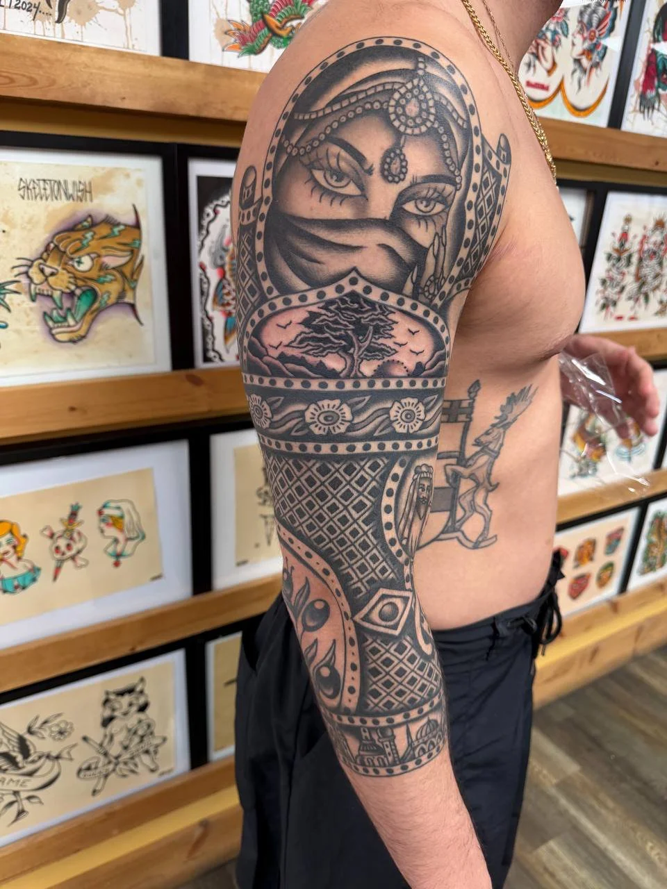 Black tattoo sleeve featuring a woman's face with detailed eyes and nose, a landscape with trees, decorative geometric patterns, and other intricate designs on a person's arm.