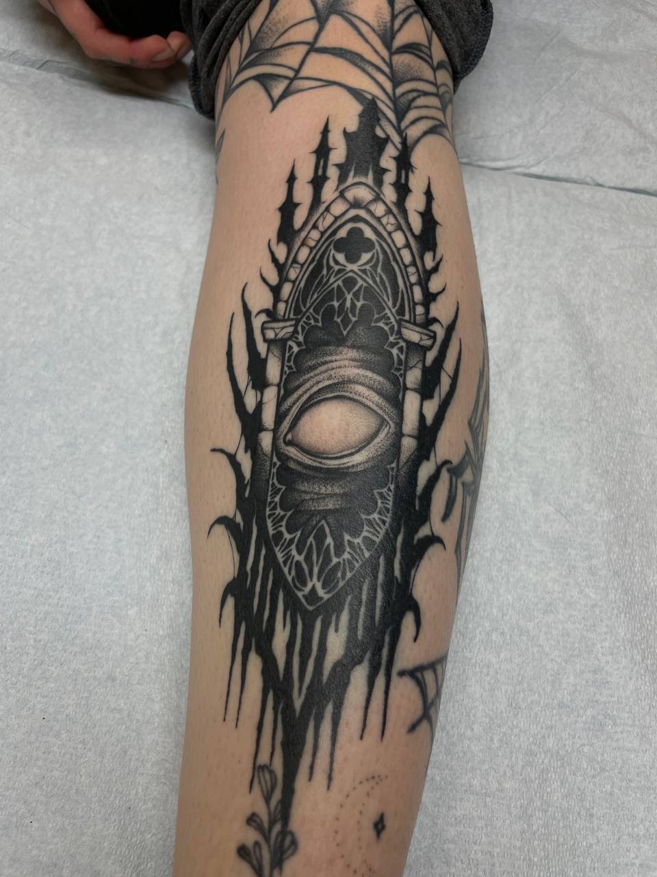 Black tattoo of a distorted eye inside a gothic-style window with sharp, jagged edges on a person's forearm.
