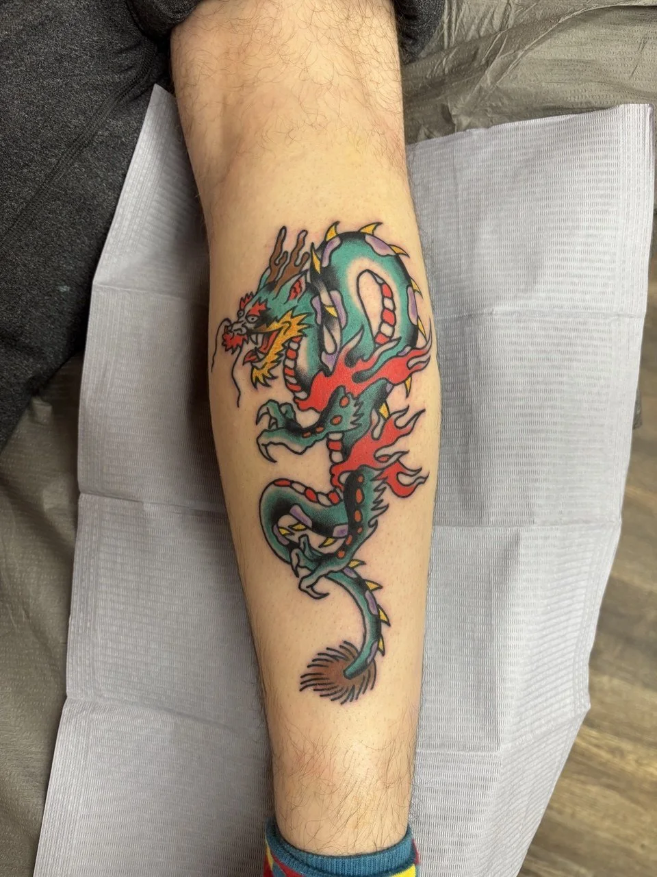 Colourful tattoo of a dragon on a person's calf, featuring a fierce face, long body, and fiery elements. Classic American traditional tattoo. 