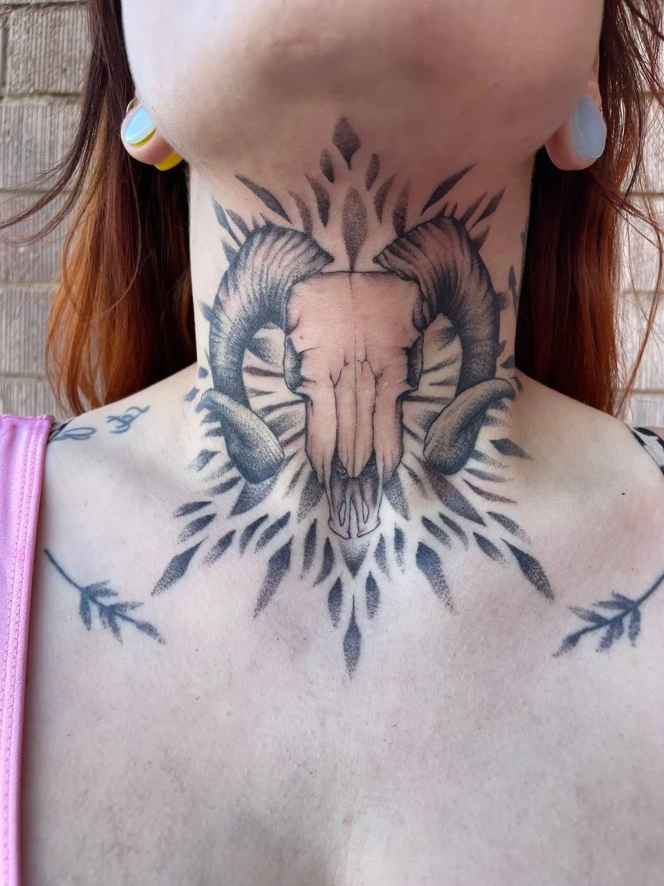 Tattoo of a ram's skull with large horns, surrounded by geometric and linework designs on the neck and upper chest.
