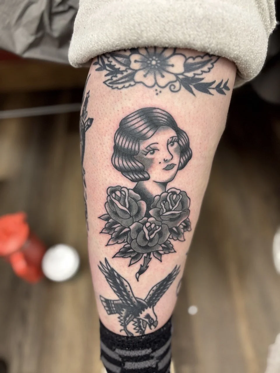 Tattoo of a woman's face with wavy hair surrounded by roses, with an eagle at the bottom. Black and grey American traditional tattoo.