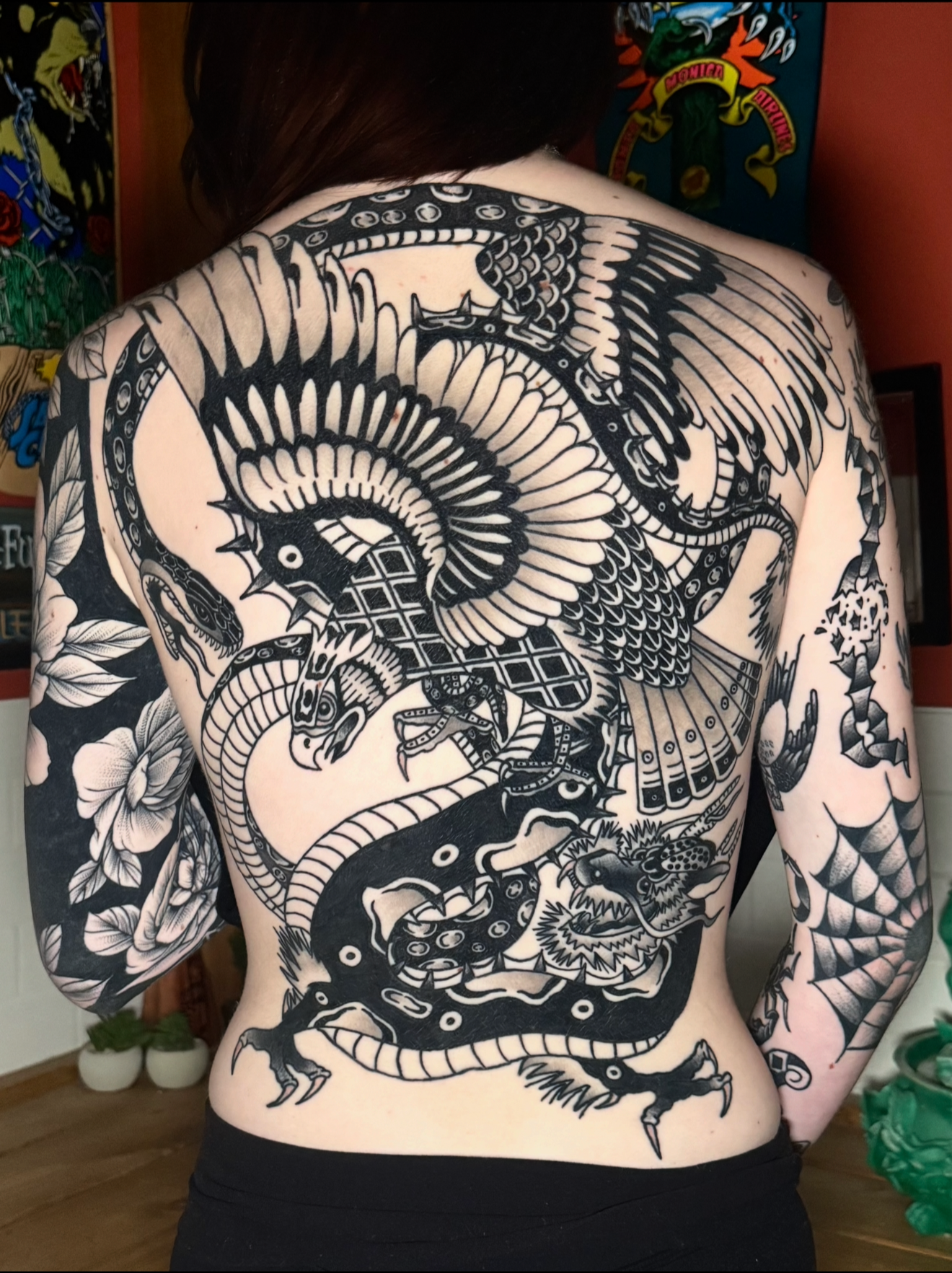 A person with a full-back black tattoo of a dragon, eagle, and snake fighting. Black and grey American traditional back piece.