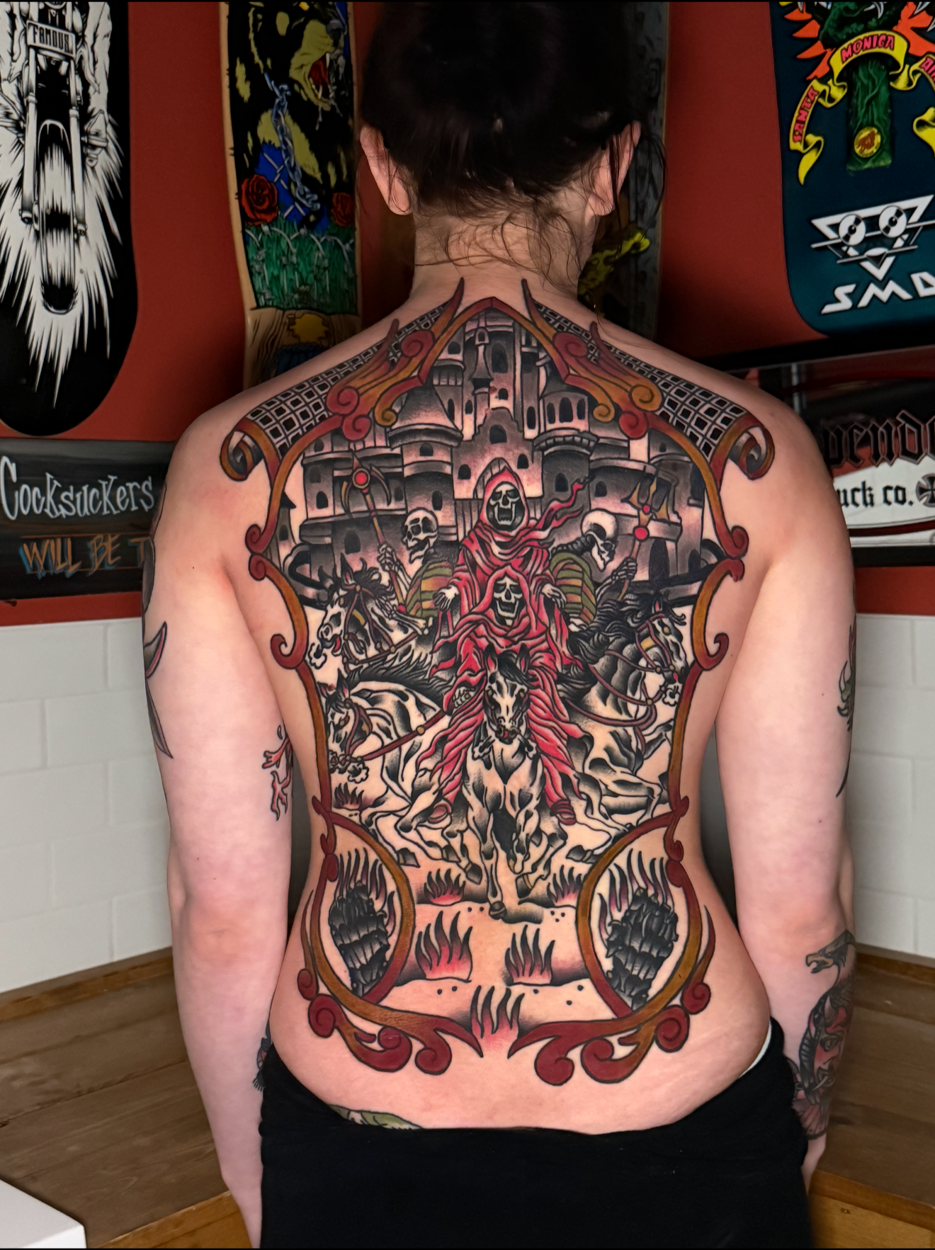 Back tattoo depicting skeletons, skulls, a castle, and flames in a dark, gothic style. Full colour American traditional back piece.
