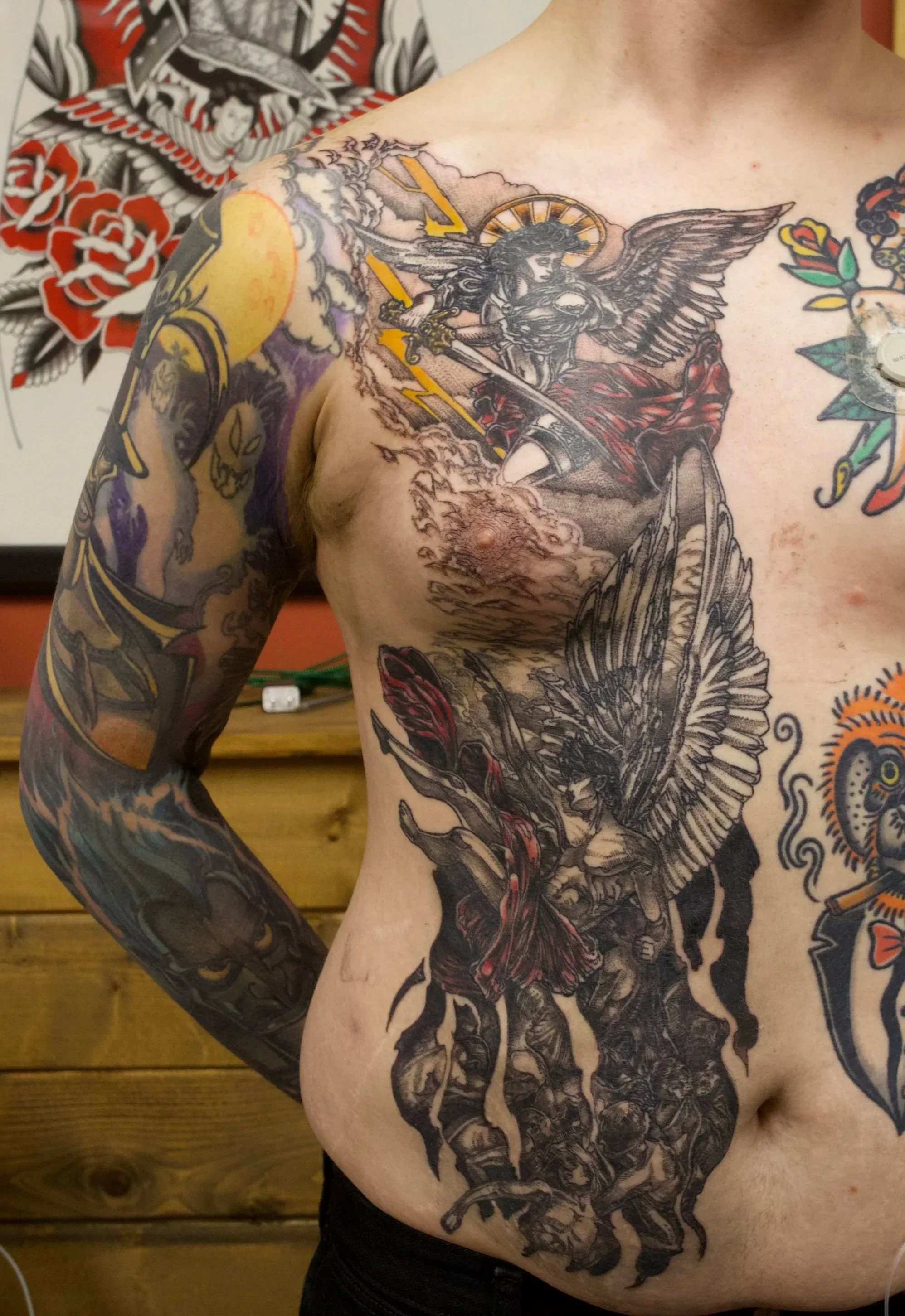 A person with extensive tattoos, including a large black and gray warrior angel with wings, holding a sword with lightning, on the chest and arm. Full colour neo traditional style tattoo.