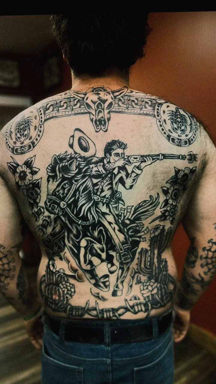 Black and white detailed tattoo of a man on horseback aiming a rifle, surrounded by flowers and decorative elements, on a person's back. Black and grey American traditional back piece tattoo.