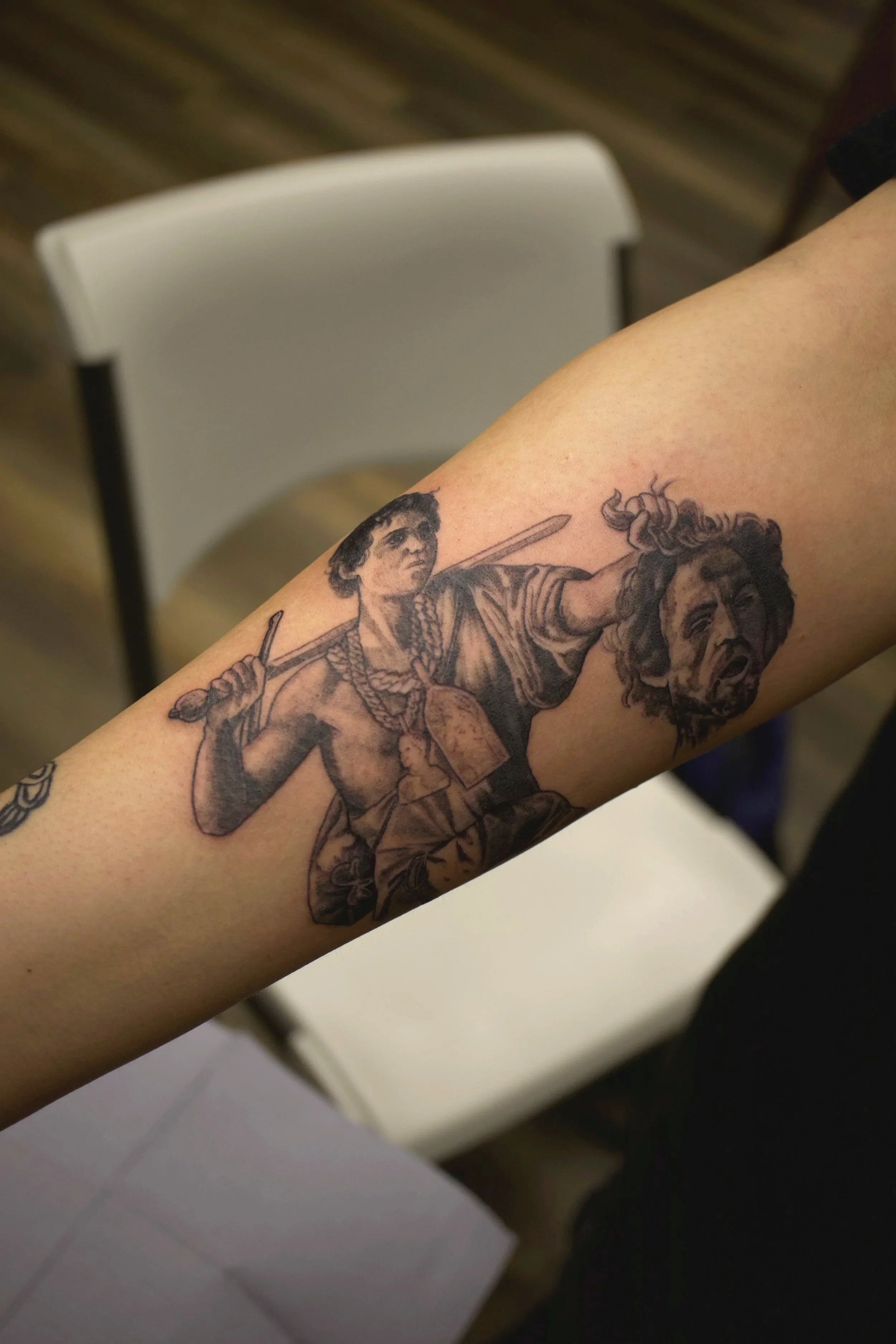 A tattoo of a man holding a sword and a decapitated head on a person's arm. Black and grey realism. David holding the head of Goliath.
