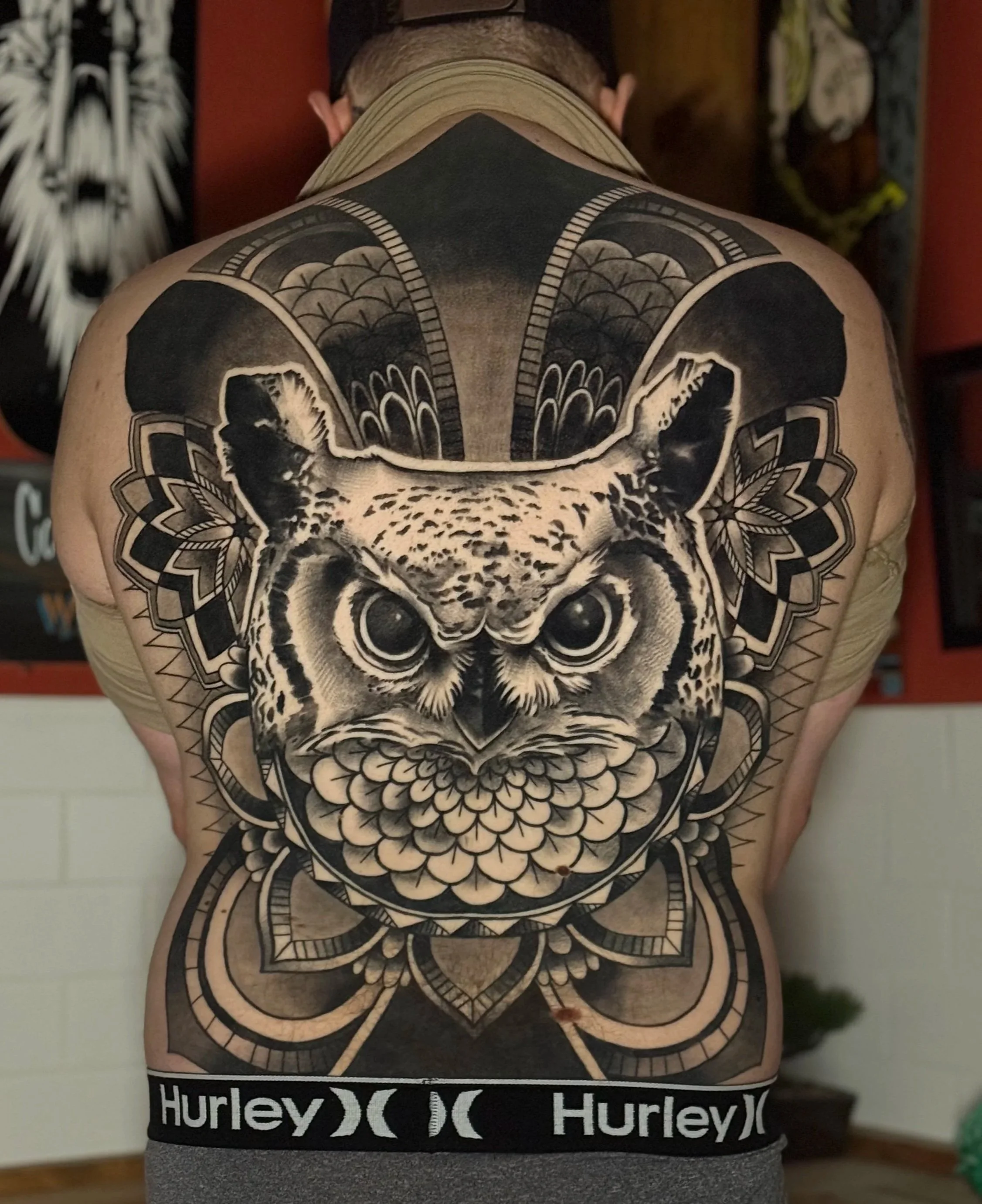Person with a large, detailed black and grey Owl tattoo on their back. The tattoo features intricate floral and geometric patterns. Black and grey realism back piece tattoo.