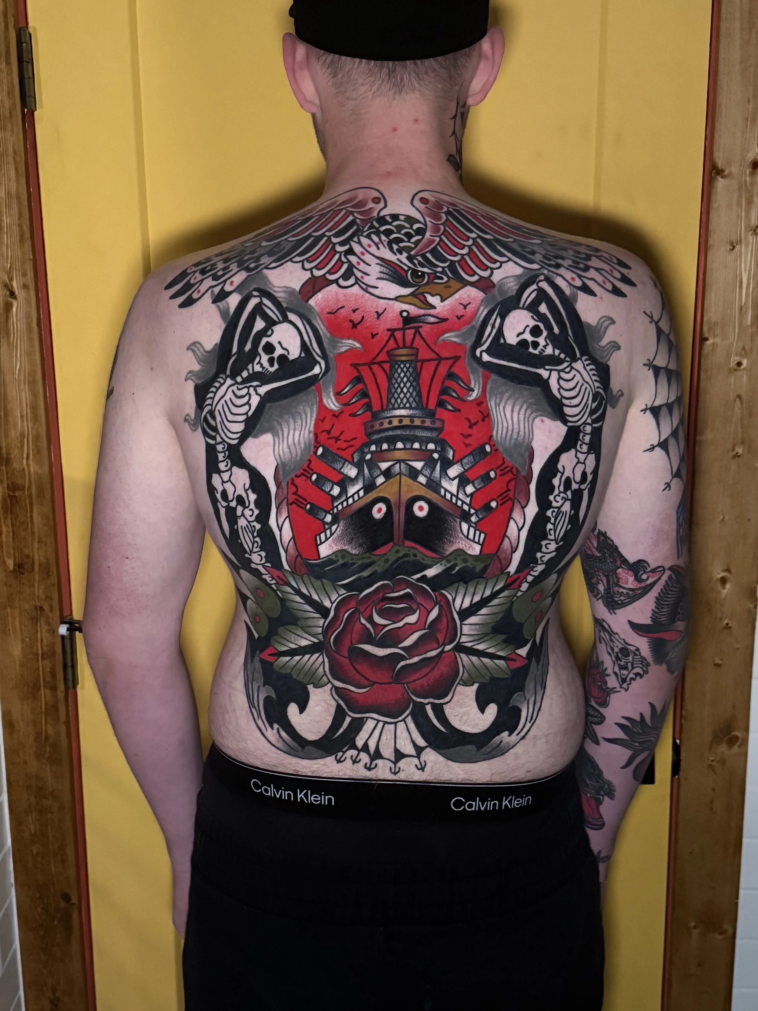 Back of a person with large tattoos depicting a traditional American traditional style, including a mermaids, skulls, a ship, and a large rose. Full colour American traditional back piece.
