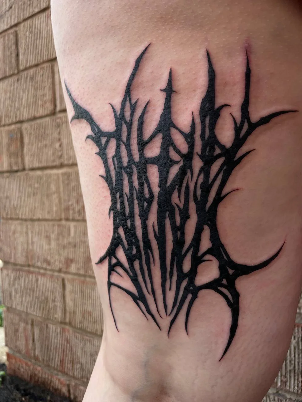 Black tribal blackwork style tattoo on person's upper arm, next to a brick wall, with sharp, jagged lines forming an abstract design.