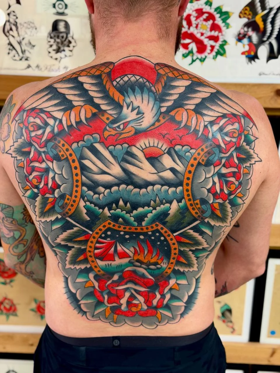 Colourful tattoo of an eagle, mountains, a sun, and a snake on a man's back. Bold American traditional tattoo back piece.