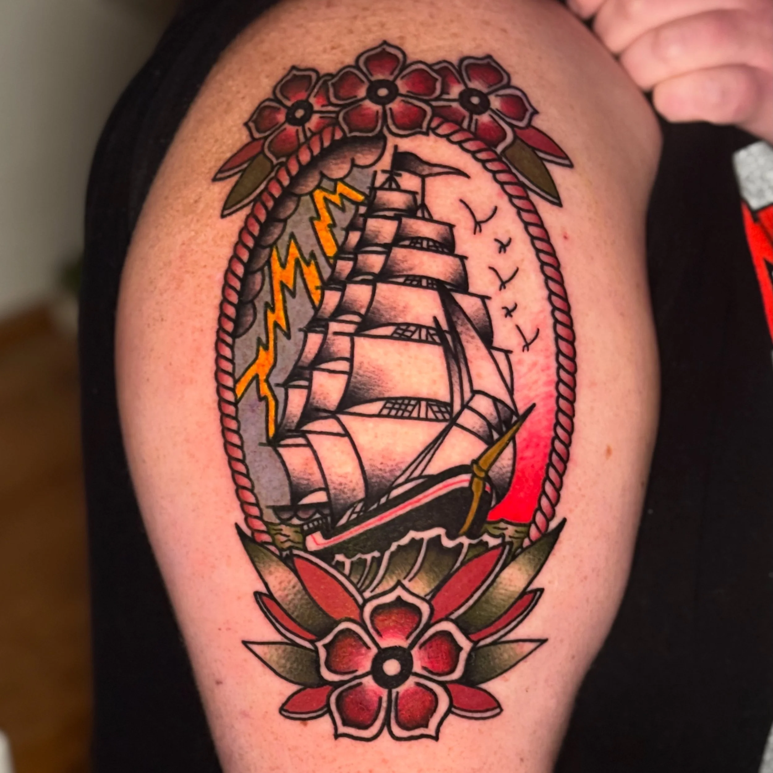 Colorful tattoo of a ship on a person's upper arm, featuring flowers, lightning bolts, birds, and clouds.