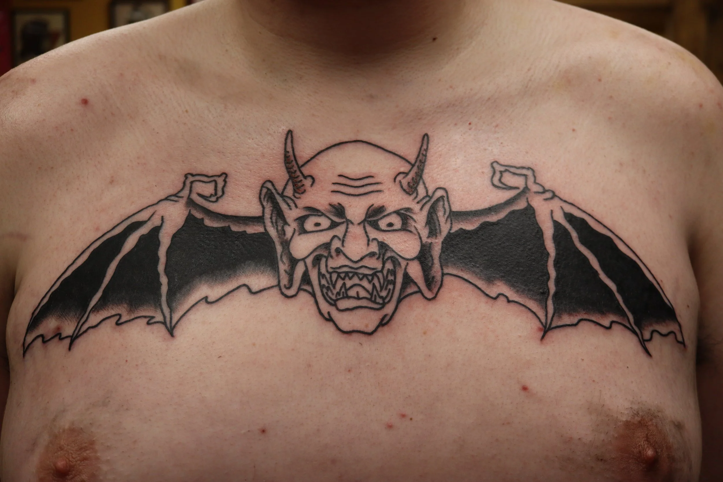 A tattoo of a fierce demon face with horns and wings on a person's chest. Black and grey American traditional tattoo.