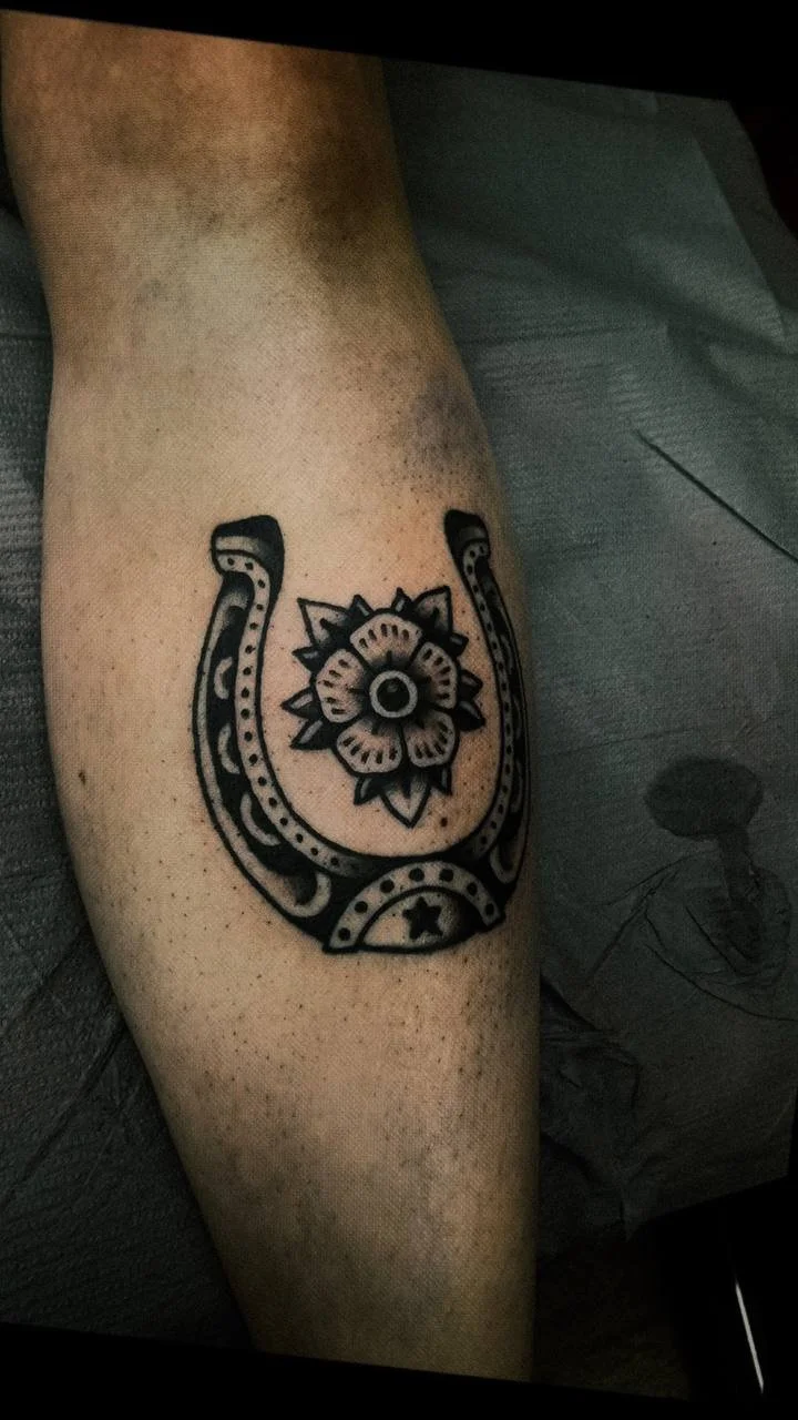 Tattoo of a horseshoe with a decorative flower in the center on a person's leg.