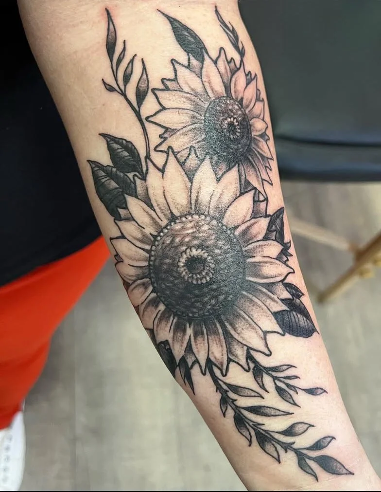 Black and gray tattoo of two sunflowers with leaves, detailed and shaded, on a person's forearm. Black and grey neo traditional tattoo.