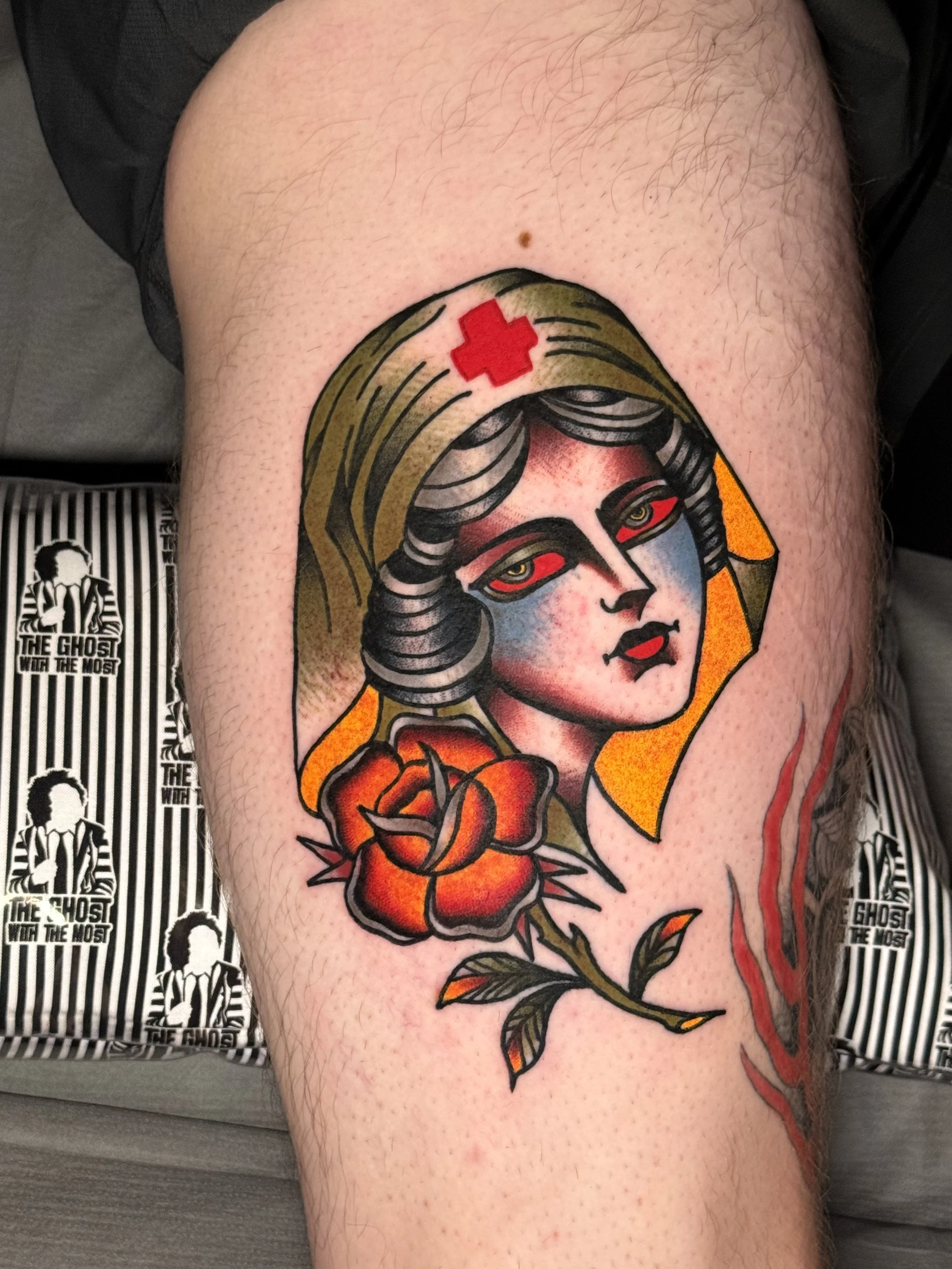 Tattoo of a nurse woman with dark hair and makeup, wearing a yellow hood, with a red cross above her head, holding a red rose on her shoulder.
