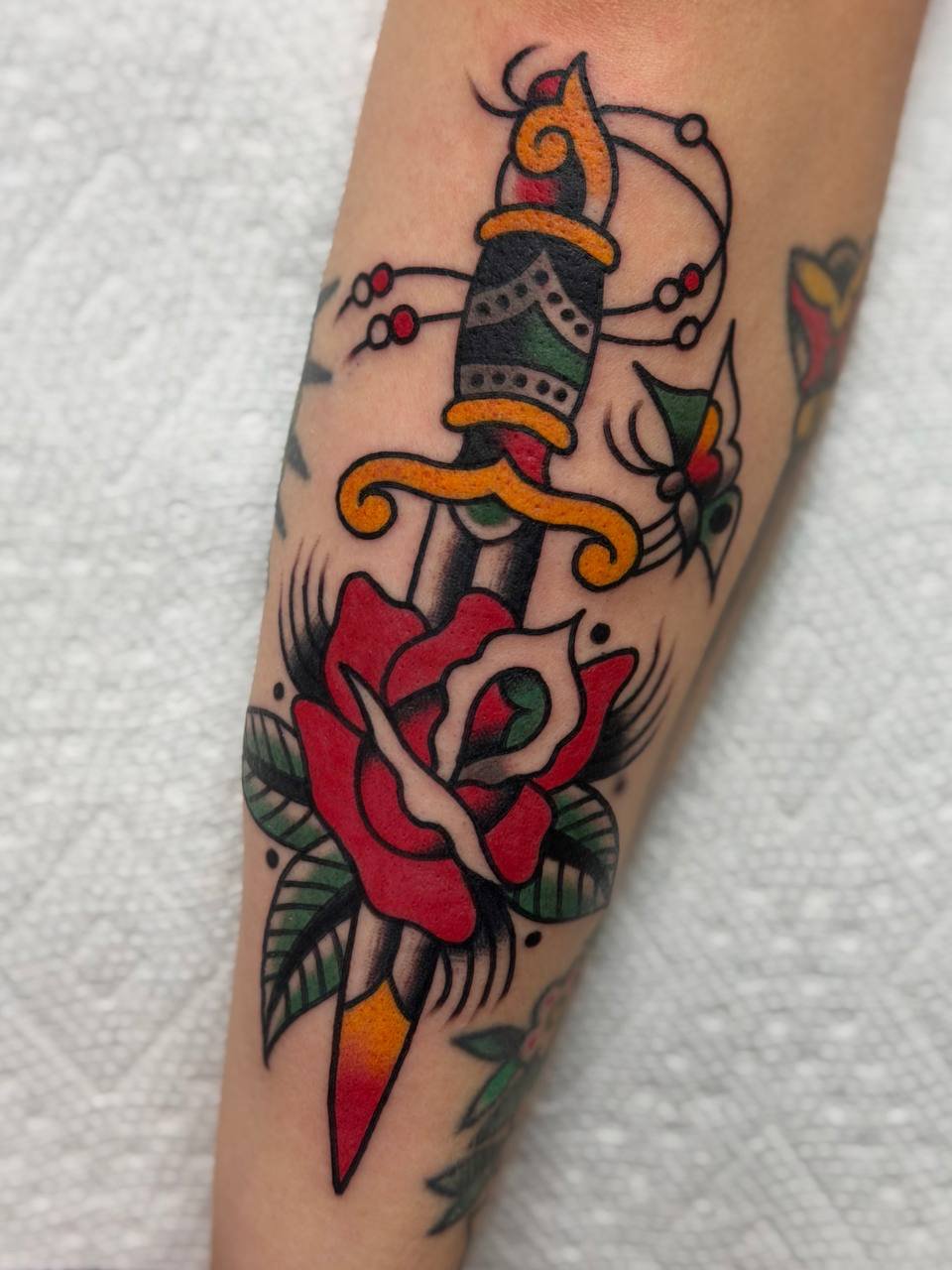 Tattoo of a dagger piercing a rose with leaves, in traditional style with bold lines and bright colors.