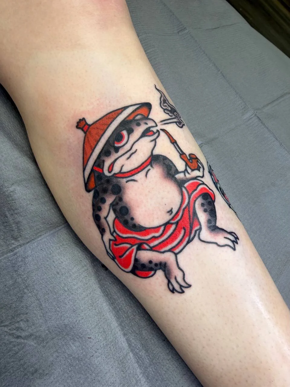 Tattoo of a frog wearing a hat and smoking a pipe, sitting with legs crossed and wrapped in a red cloth. Bold Japanese traditional tattoo.