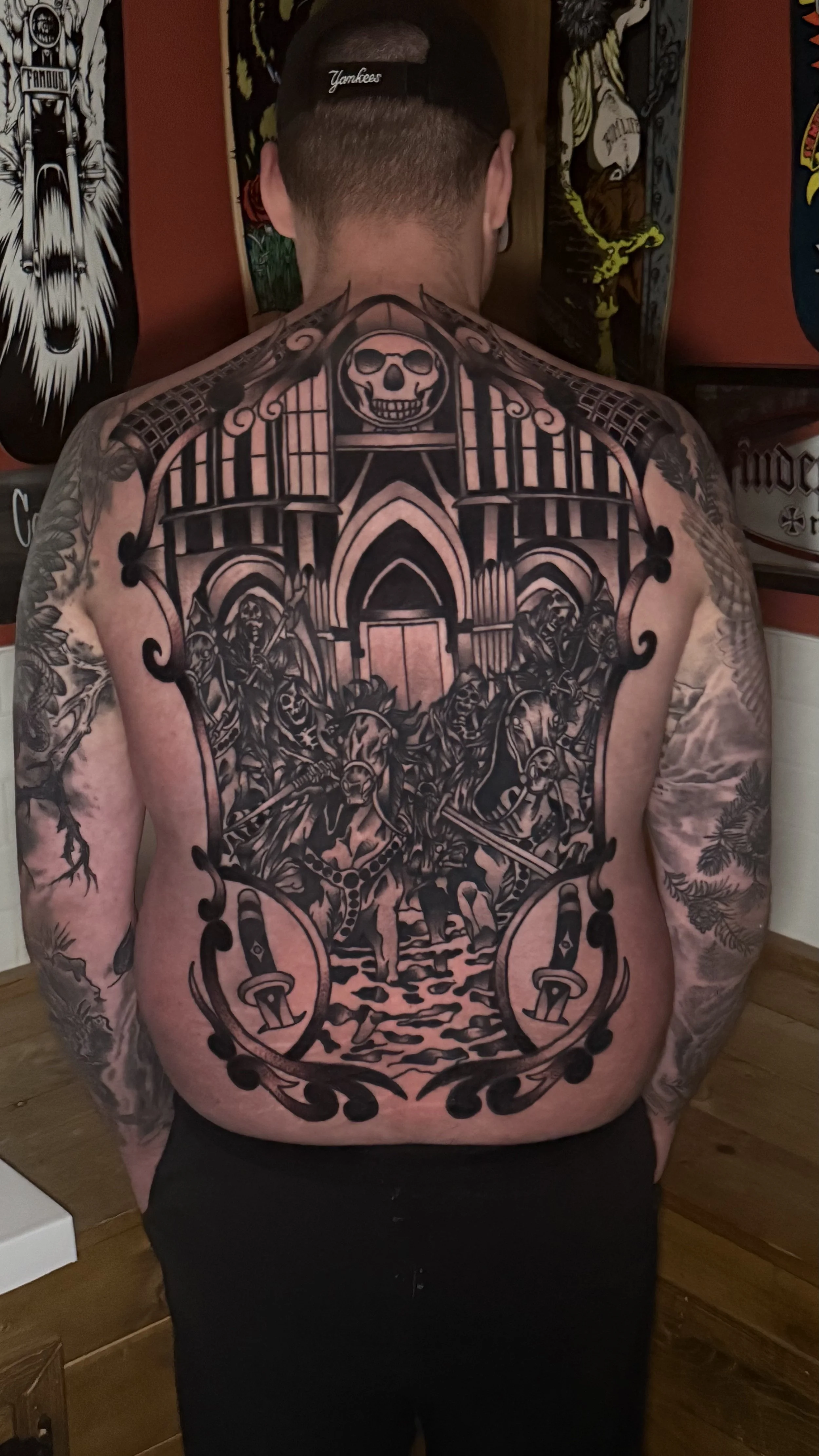 Back view of a man with extensive black and grey tattoo covering his back and arms, featuring a gothic church, skeletons, and a skull at the top. Four horsemen of the Apocalypse. Black and grey American traditional back piece.