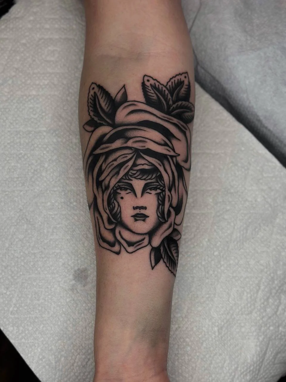 Black ink tattoo of a woman's face with a headscarf and leaves on the arm.